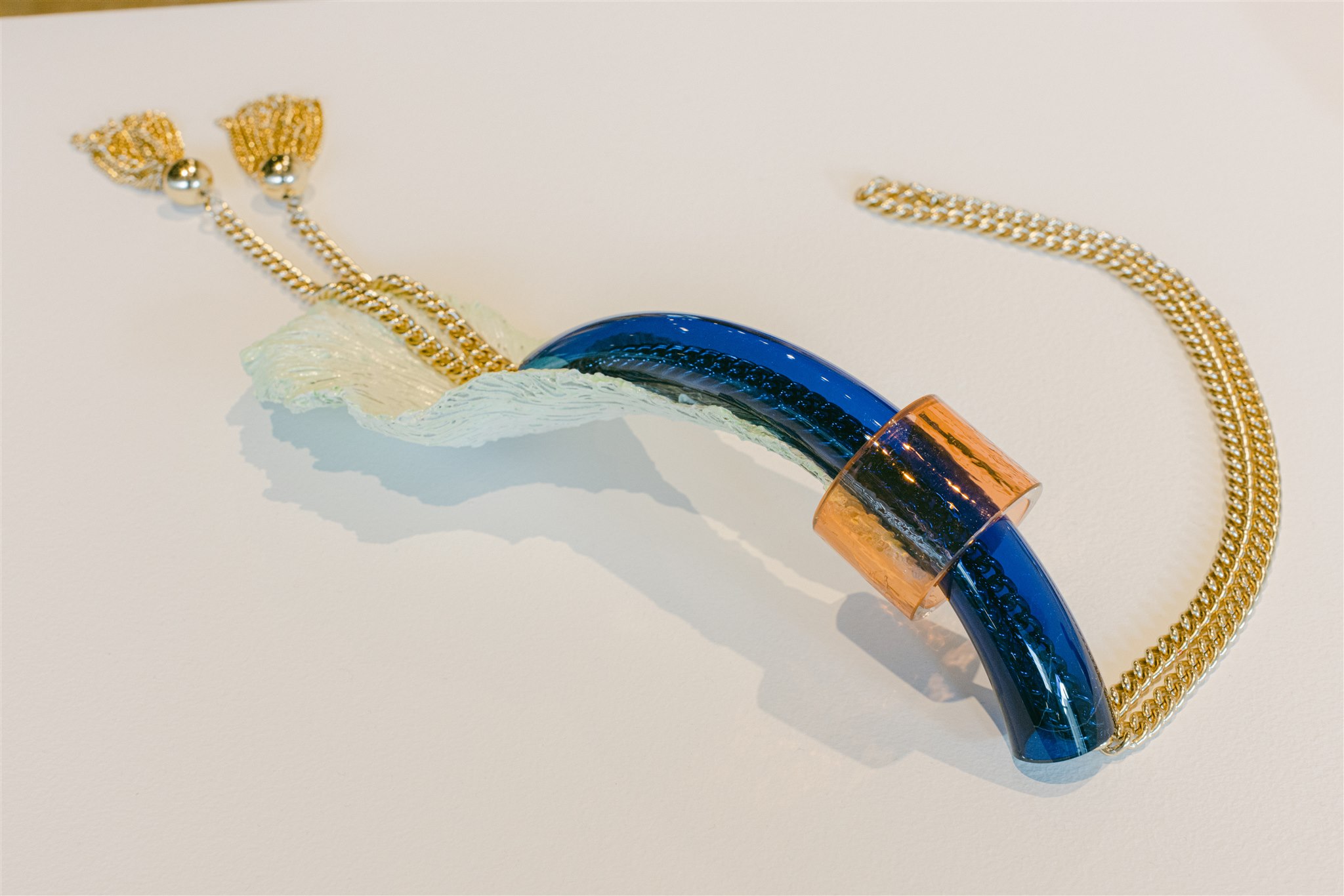 Colorful art necklace made of gold chain, glass, and plastic on white pedestal