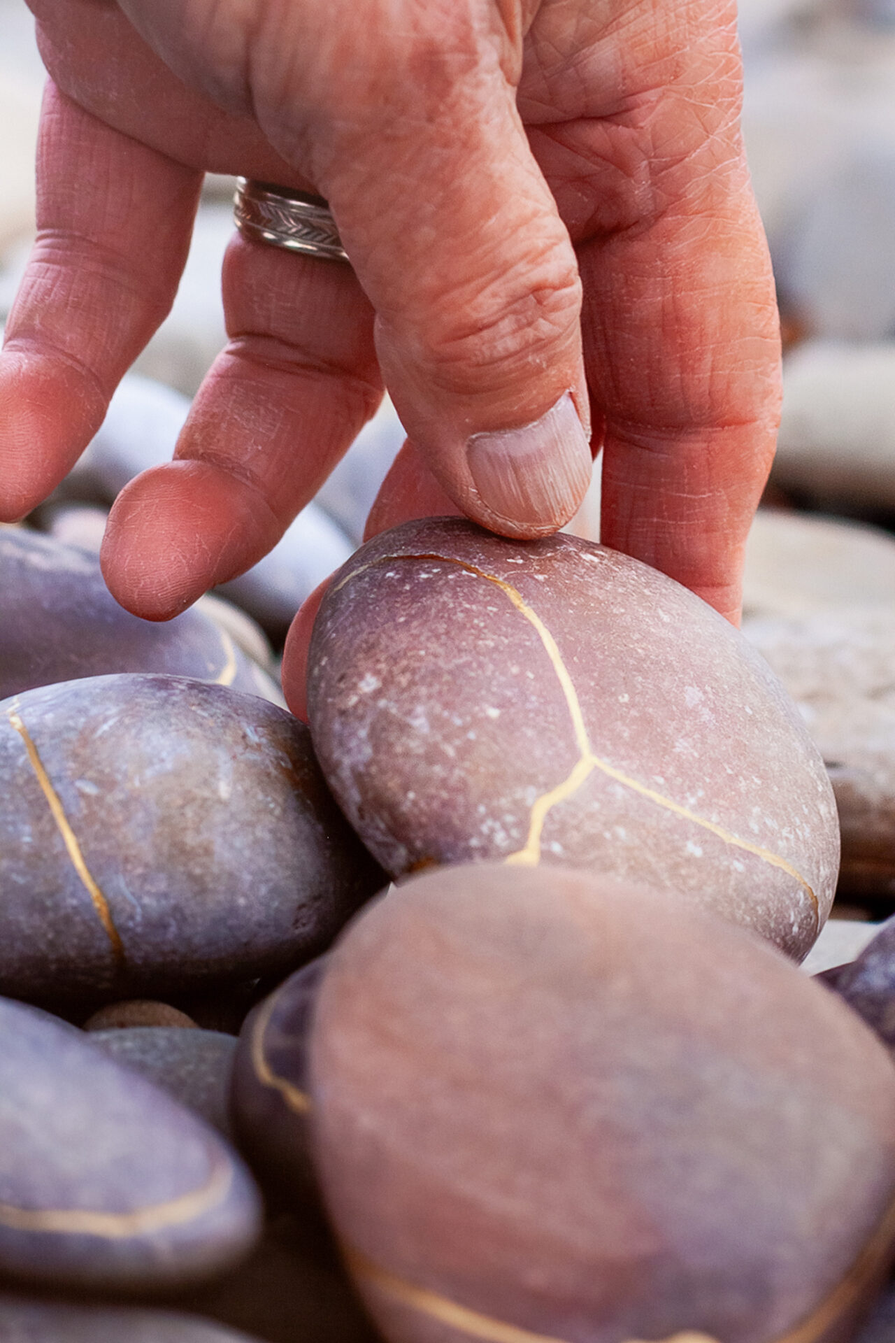 A close-up of a hand reaching for a smooth, rounded stone among several others, showcasing various shades of purple and grey with distinct golden lines running through them. The hand appears to have a silver ring and a natural nail finish, emphasizing the tactile interaction with the stones.