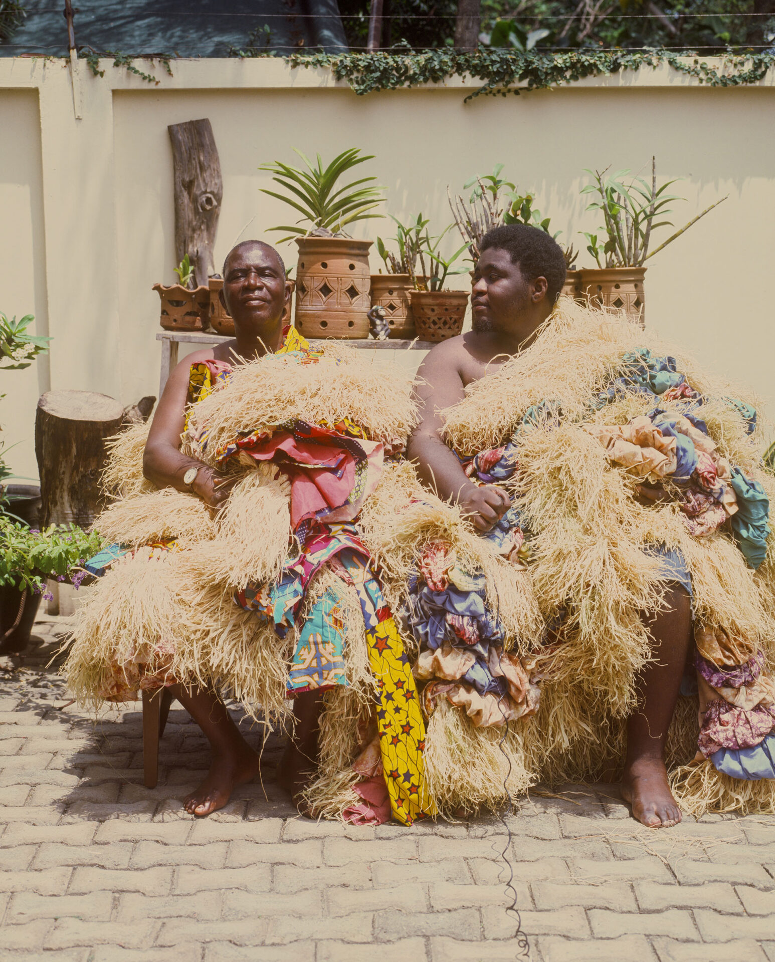 Two men sitting on a bench wearing garments made from raffia and multicoloured fabrics.