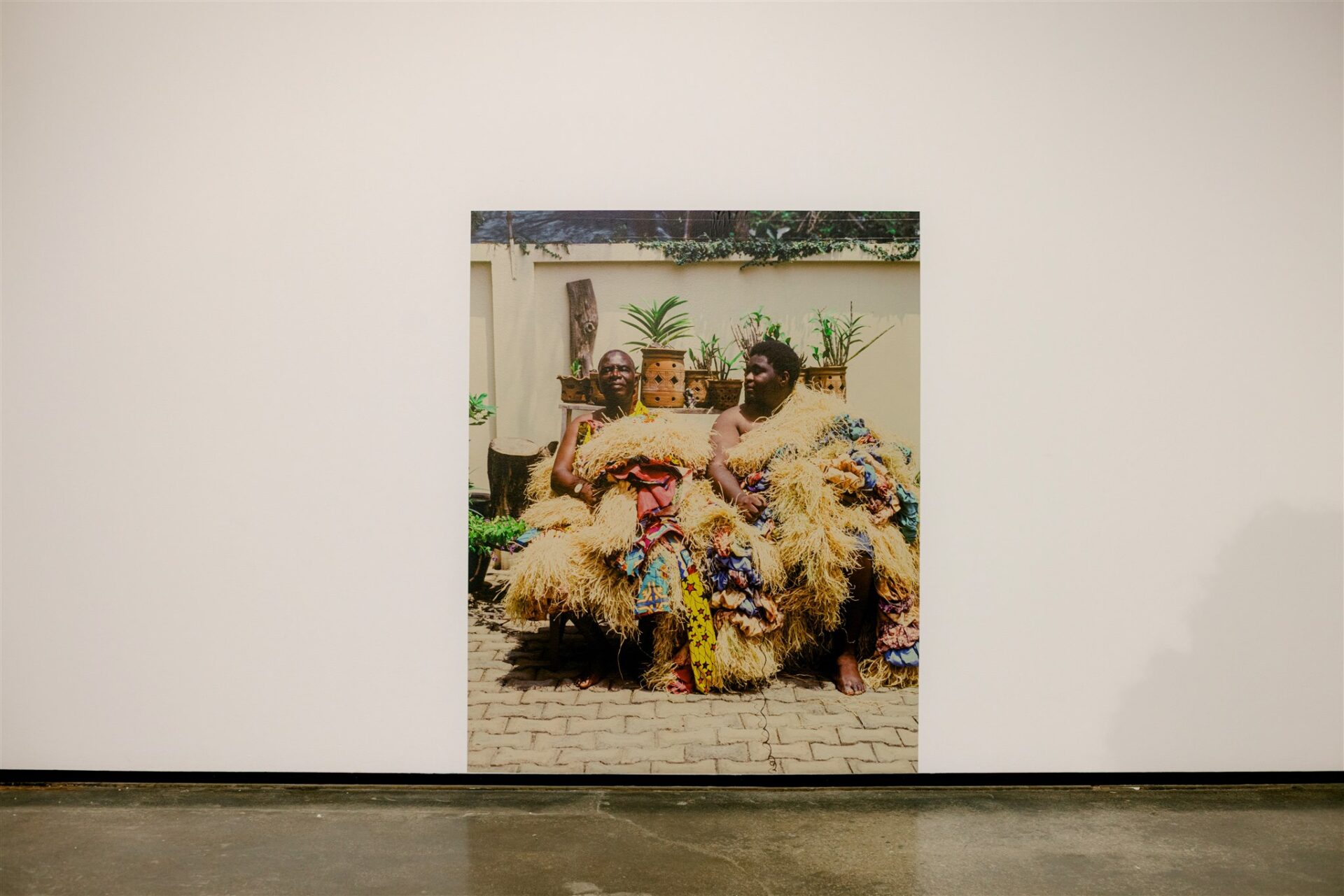 Two people in colorful straw outfits seated outdoors, surrounded by plants