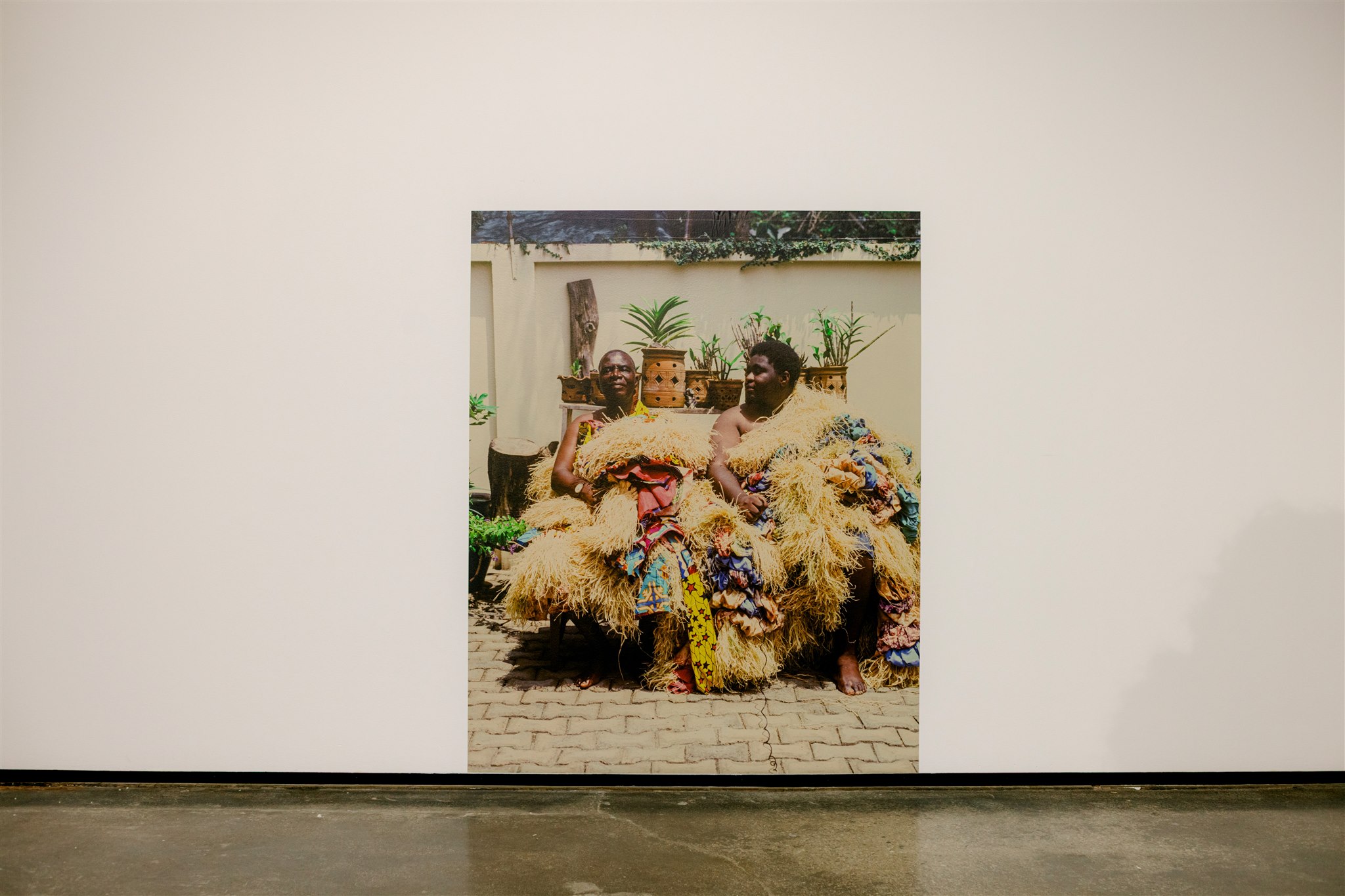 Two people in colorful straw outfits seated outdoors, surrounded by plants