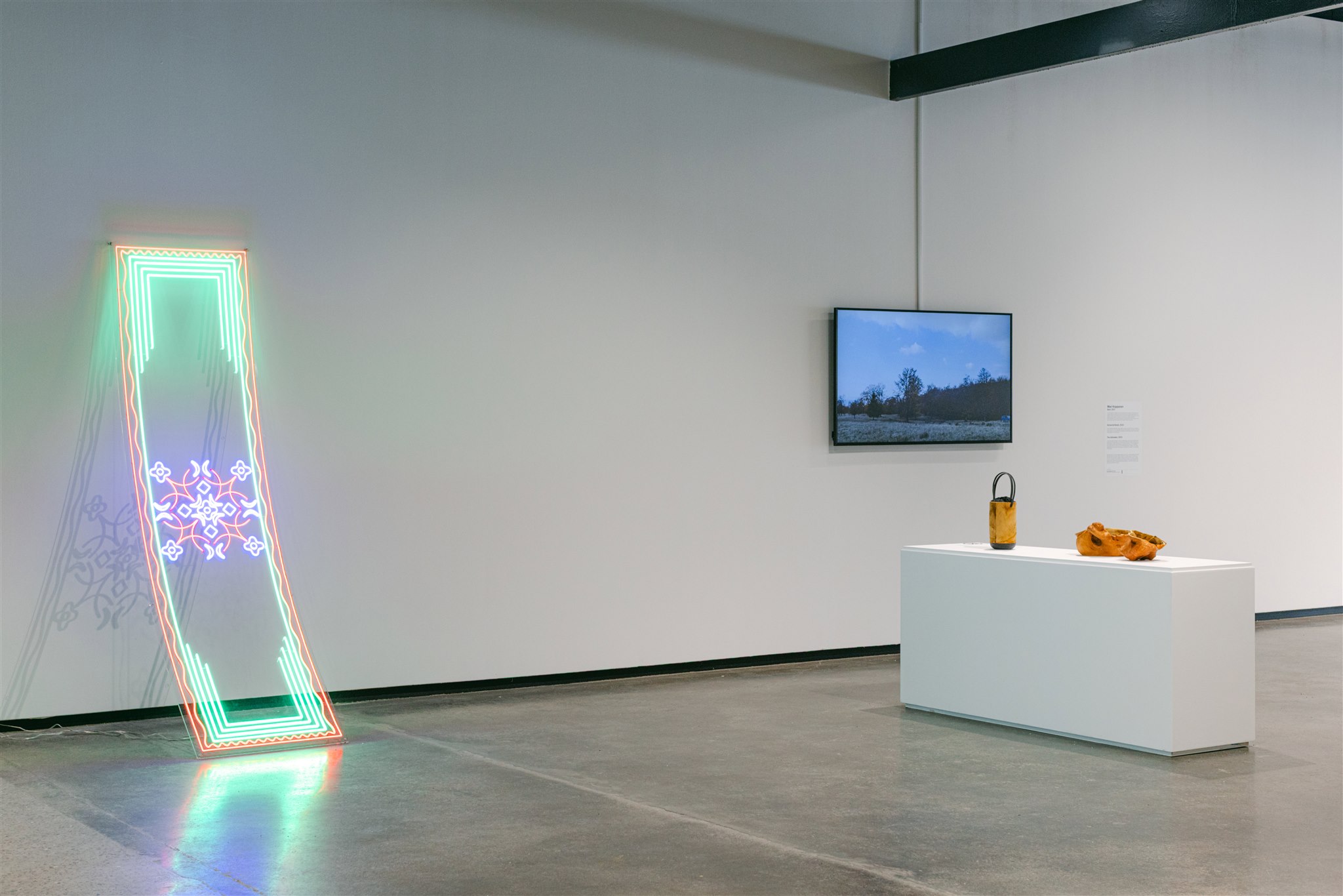 Neon wall piece, video screen, and wooden vessels on a white pedestal