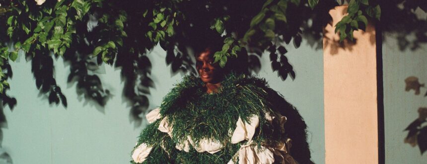 Person in a foliage-like costume blending into greenery against a teal wall with white blossoms overhead.