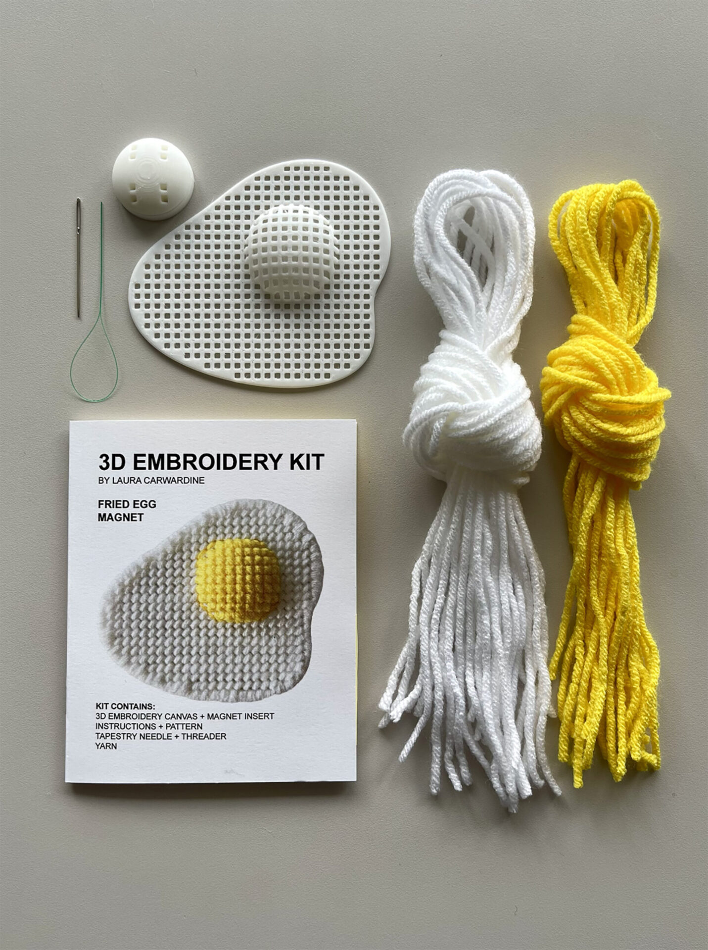 Embroidery Kit Contents on a Tabletop (showing a booklet, egg canvas, needle, and yarn)