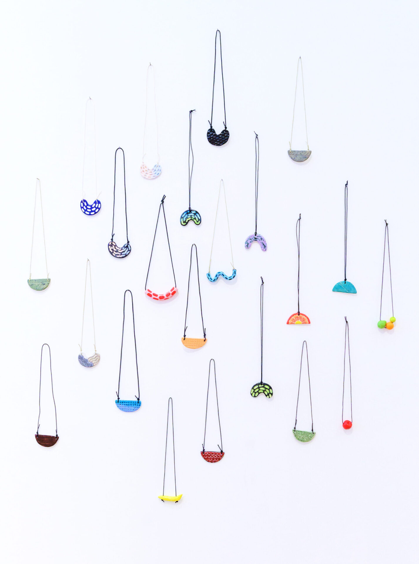 Many necklaces hang in a cluster formation on a wall. The necklaces are colourful ceramic pendants strung on cords.
