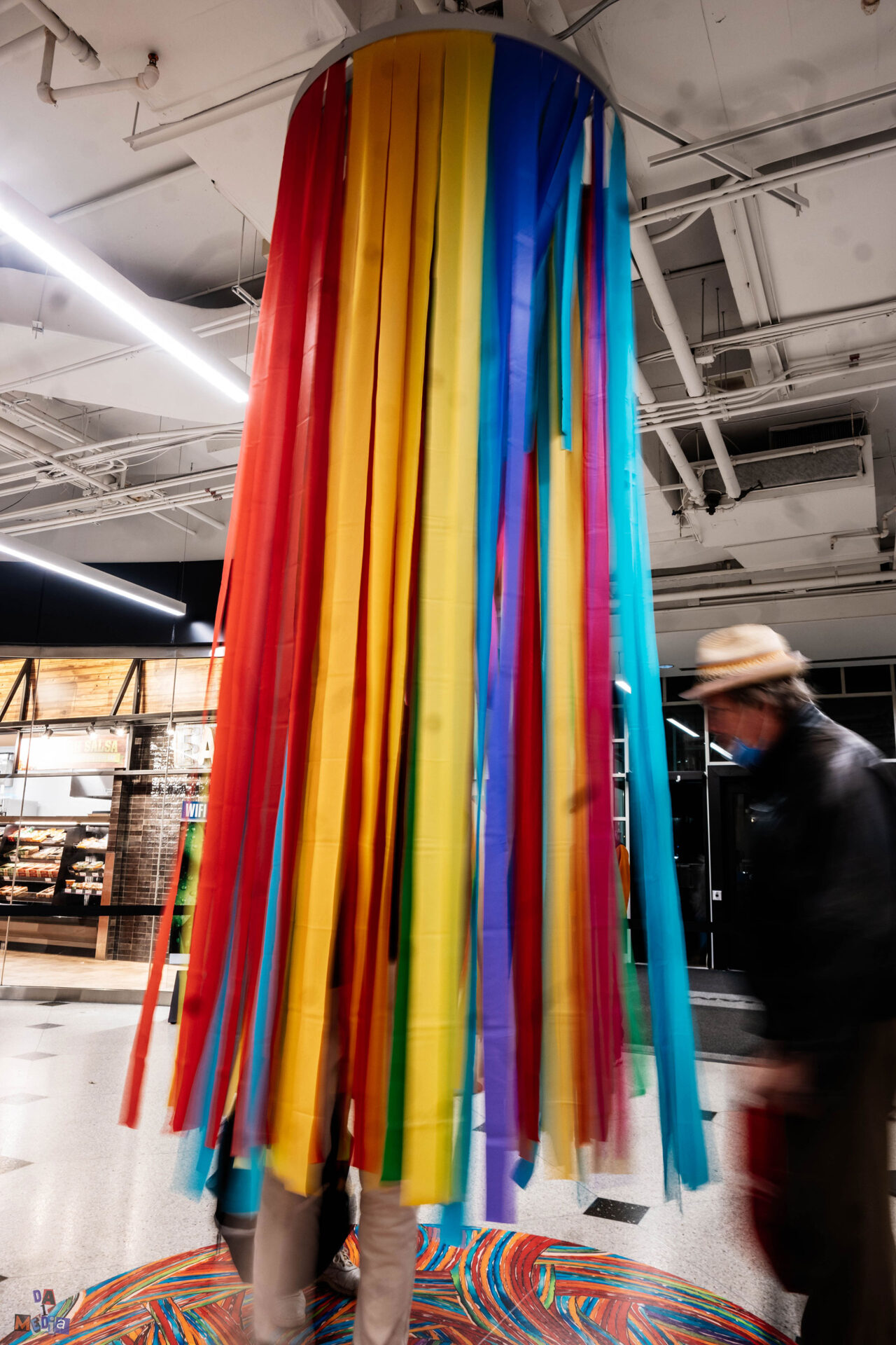 a man with a hat enters the cluster of multicoloured ribbons coming from ceiling