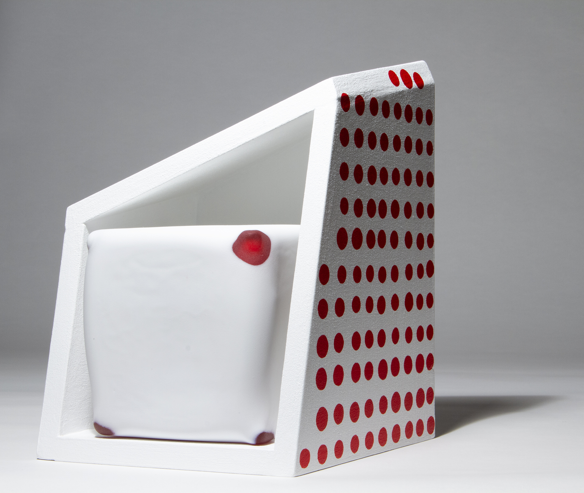 A matte white and red glass cube inside a white wood artwork with painted red dots on the right side.