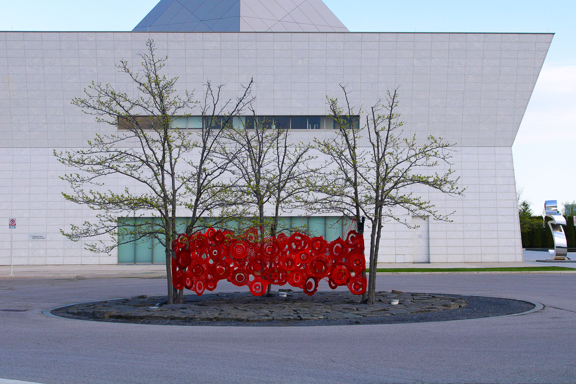 Red art installation created with red round weavings connected by crochet suspended around three trees in a triangle.  Aga Khan Art Museum.  Toronto ON