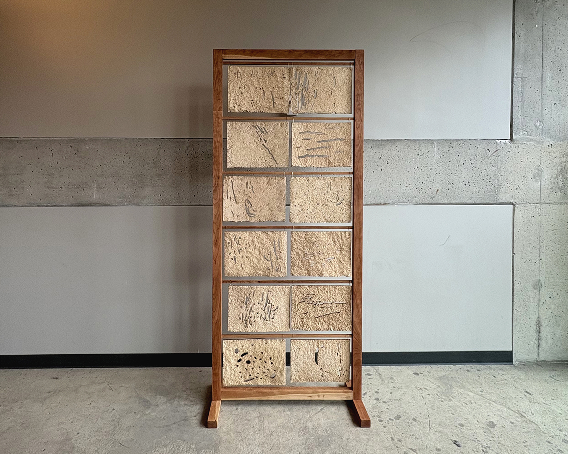 Space divider made of cherry wood and sawdust panels standing in a grey, concrete room with natural light coming from the right. Frame is warm toned wood, sawdust panels are beige and have cut out patterns from ambrosia beetle boreholes. Space divider is 6ft tall and 2.5 ft wide. 