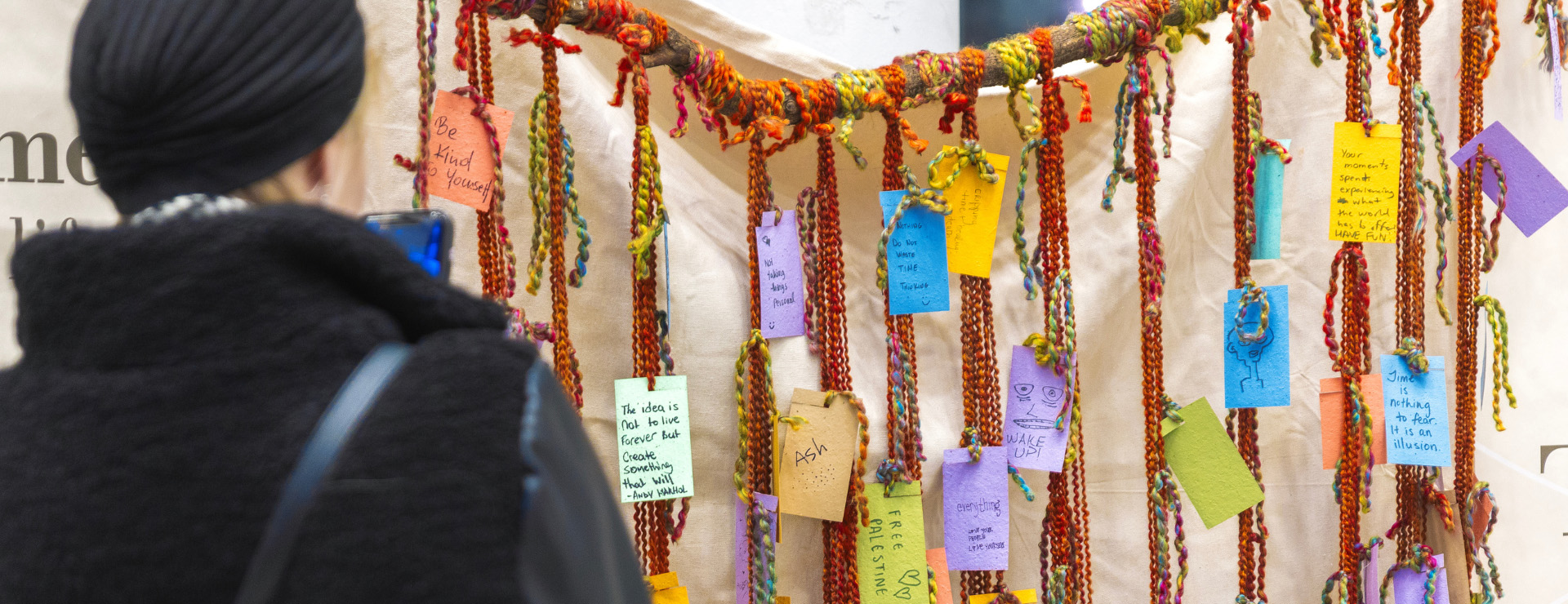 An attendee in a black knit hat stands with their back to the camera, looking at an interactive art installation. Colorful, hand-written tags are tied with vibrant, yarn-like threads to a horizontal branch. A large banner with text is visible in the background.
