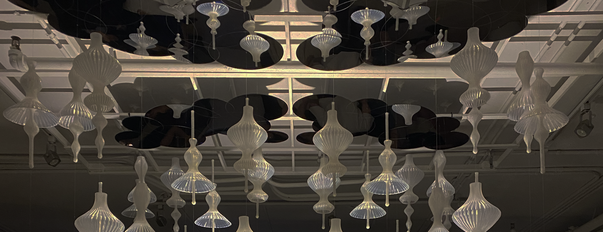 The image shows an installation of delicate, sculptural ornaments hanging from the ceiling. The forms are elegant and organic—like elongated flower buds or seed pods—crafted from translucent, ribbed material that softly glows under ambient light. Above them, glossy mirrored discs are suspended, reflecting the light and creating a layered, cloud-like canopy. The overall atmosphere feels calm, sophisticated, and ethereal, as if the ornaments are gently floating or blooming in midair.