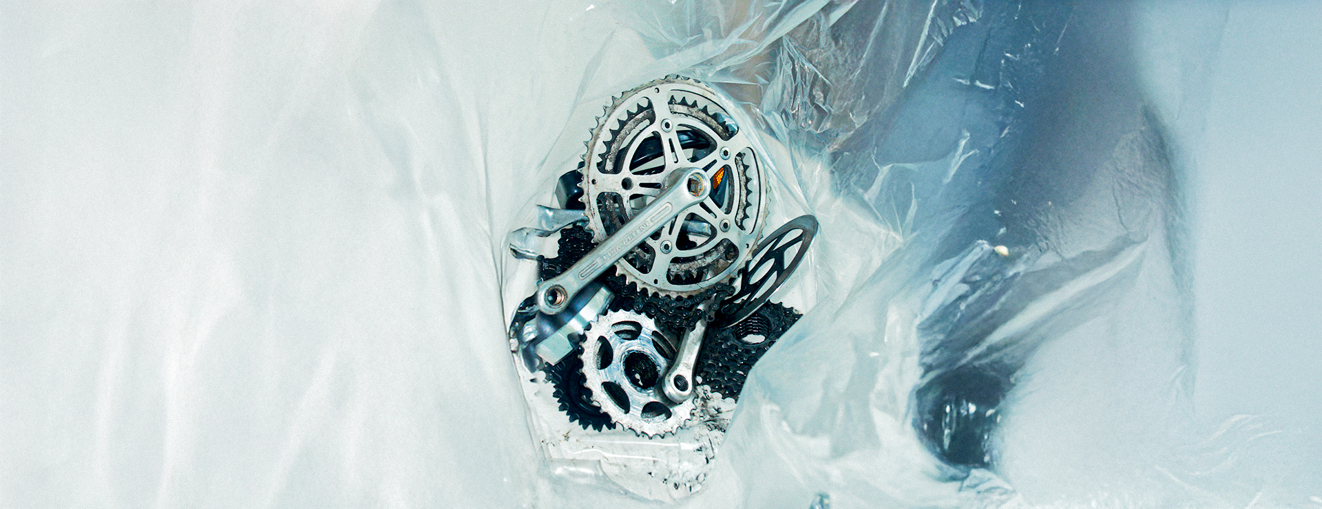 A close-up photograph of a pile of silver bicycle gears and cranksets wrapped in translucent plastic.