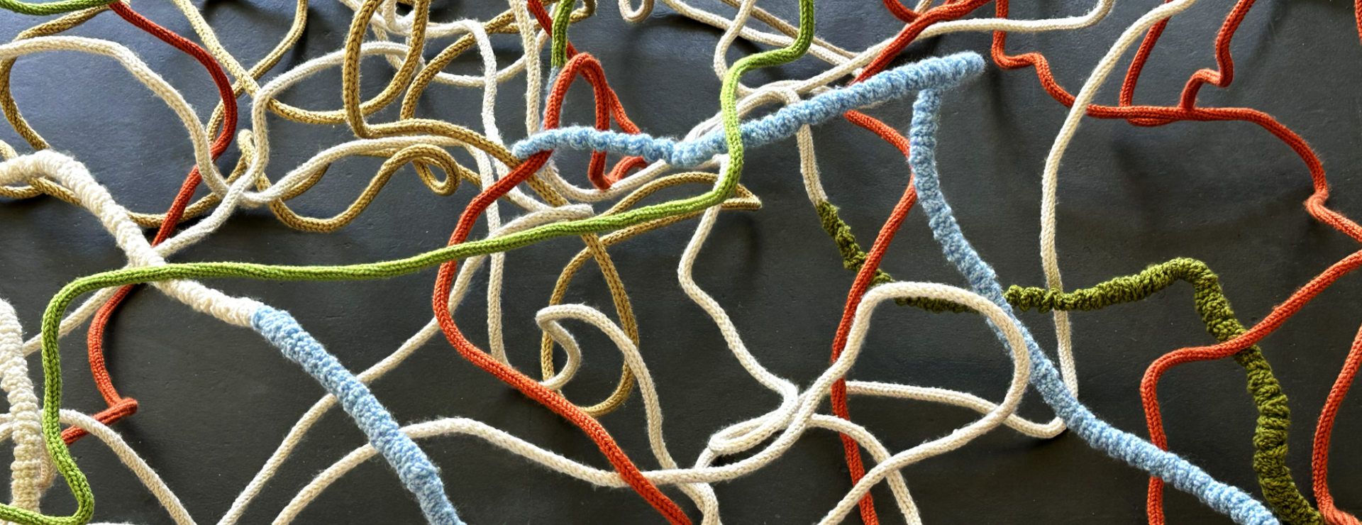 Sprawling soft sculpture made of knitted and textured yarn cords (orange, green, light blue, mustard yellow) on a dark background.