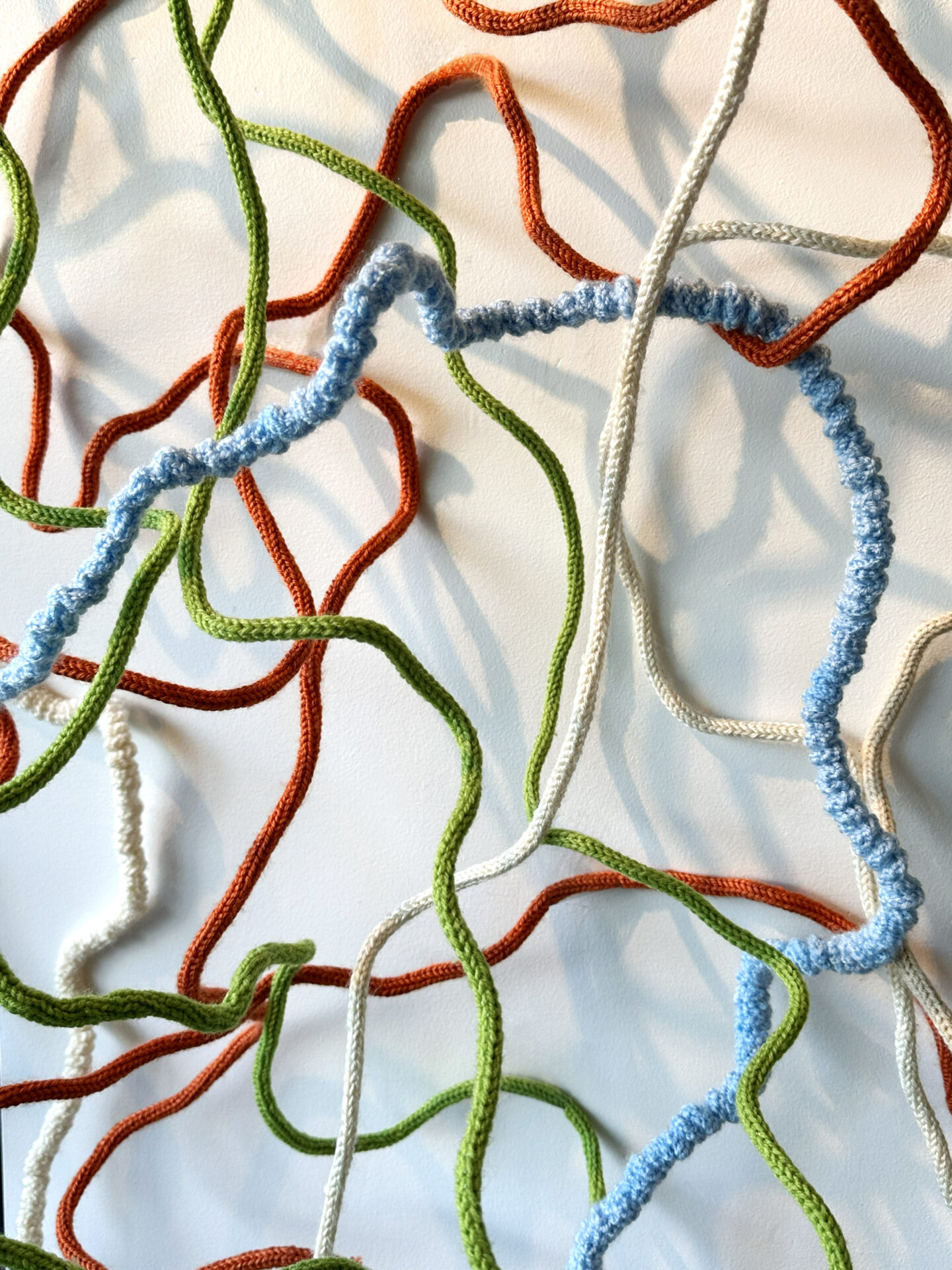 Sprawling soft sculpture made of knitted and textured yarn cords (orange, green, light blue, mustard yellow) on a light background.