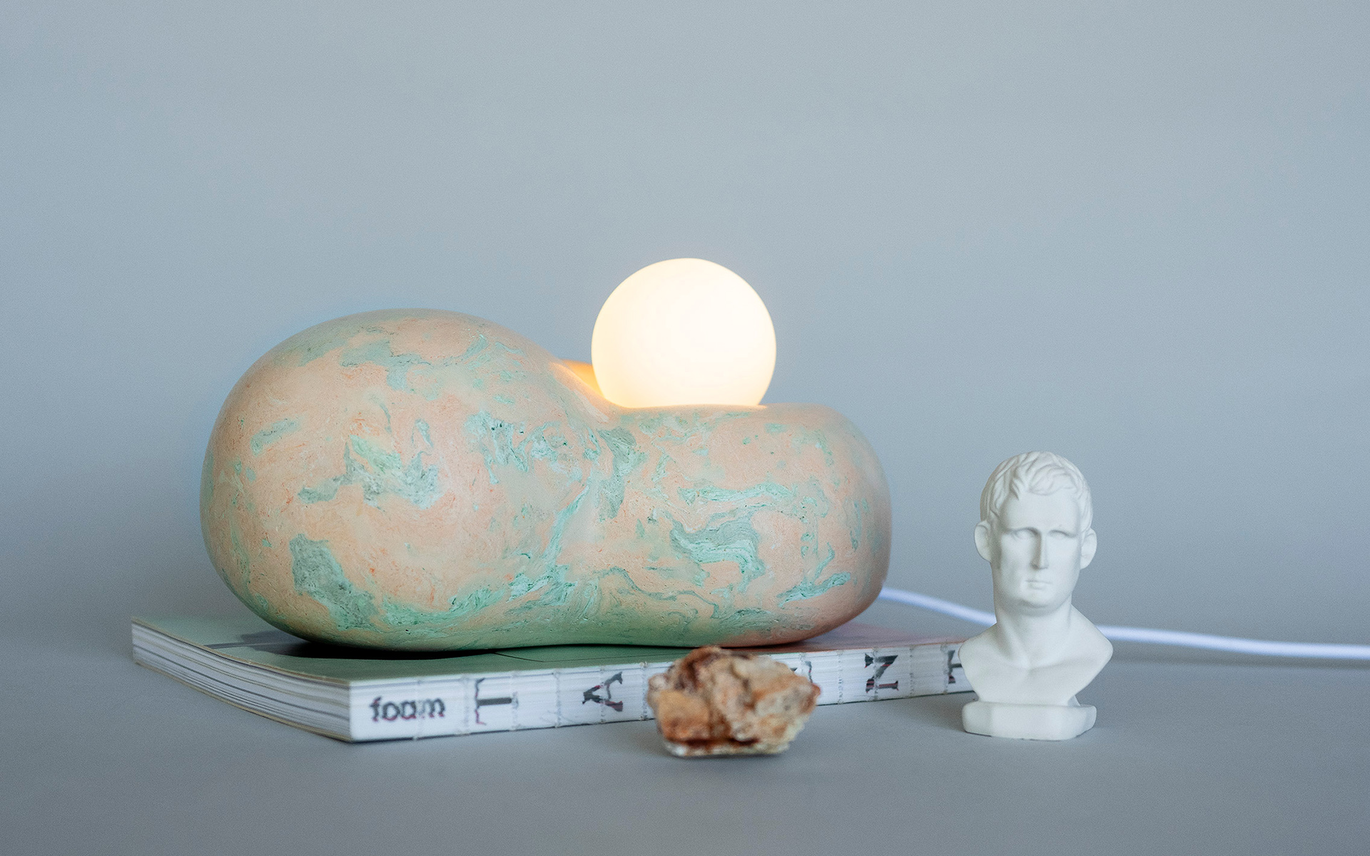 Dream Collector table lamp. An ethereal sculptural lighting piece that captures dreams in its organic embrace. Two fluid forms nestle together with luminous spheres that create a mesmerizing interplay of light and shadow. Each Dream Collector features unique scagliola patterns in soft coral and mint tones, transforming any space into a sanctuary of gentle illumination.