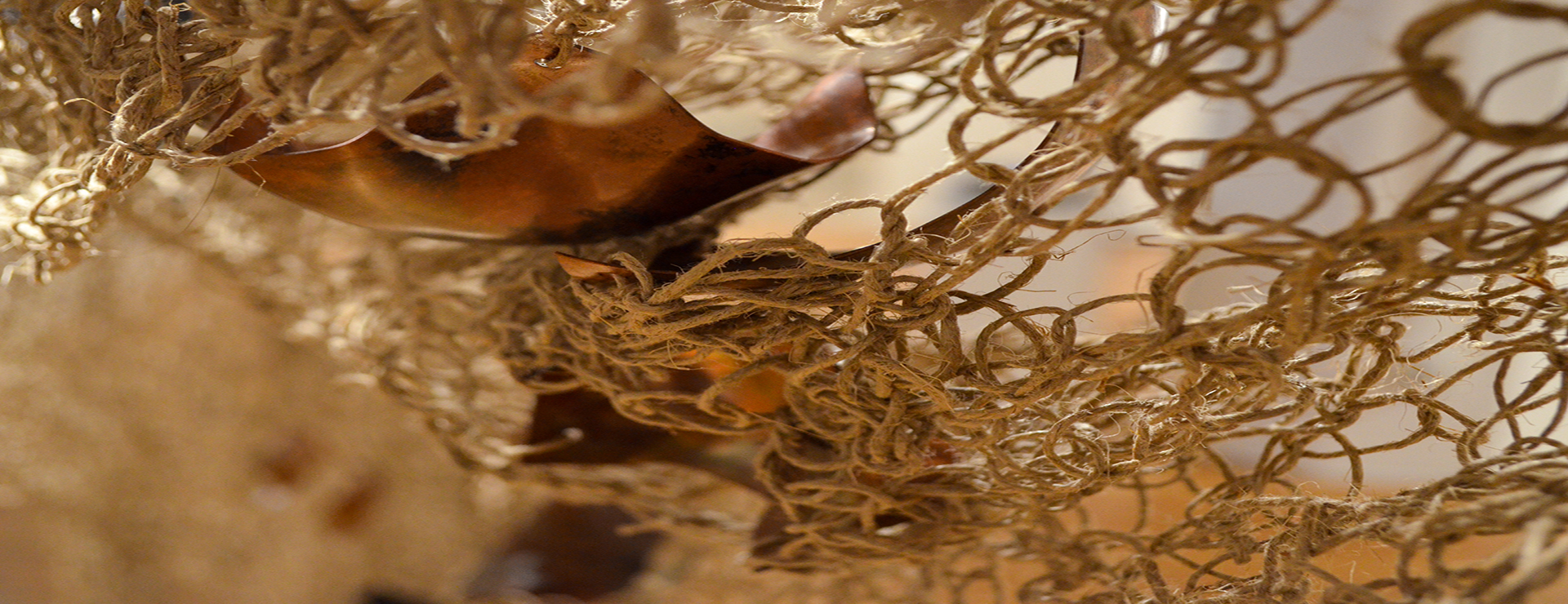 The image depicts a handwoven textile mesh made from hemp that contains fragments of organically shaped copper.