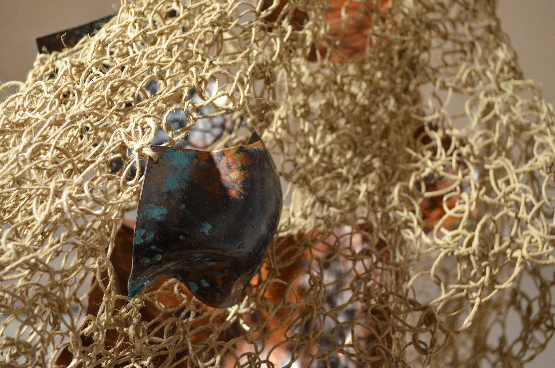 The image depicts a handwoven textile mesh made from hemp that contains fragments of organically shaped copper. 