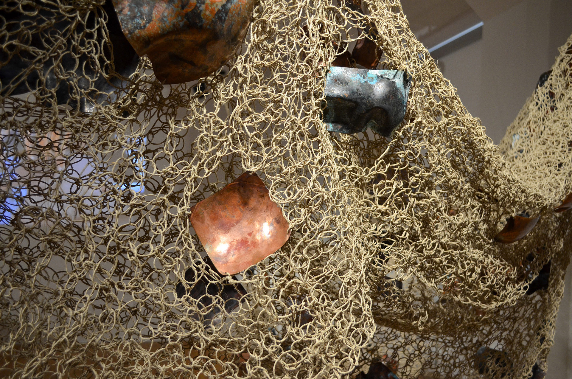 The image depicts a handwoven textile mesh made from hemp that contains fragments of organically shaped copper.