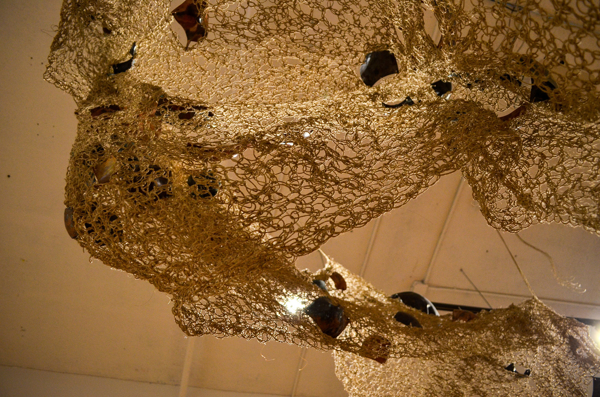 The image depicts a handwoven textile mesh made from hemp that contains fragments of organically shaped copper.