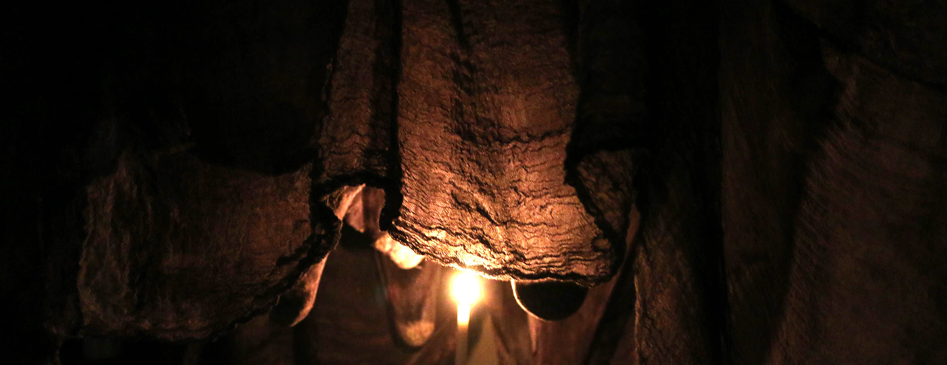 The close-up image shows a section of a cloth hanging from the ceiling, pulled downward by multiple small sand pockets. A single light source illuminates the center, revealing delicate folds and textures. The lighting creates dramatic depth and layered shadows, emphasizing the fabric’s tension and subtle movement.