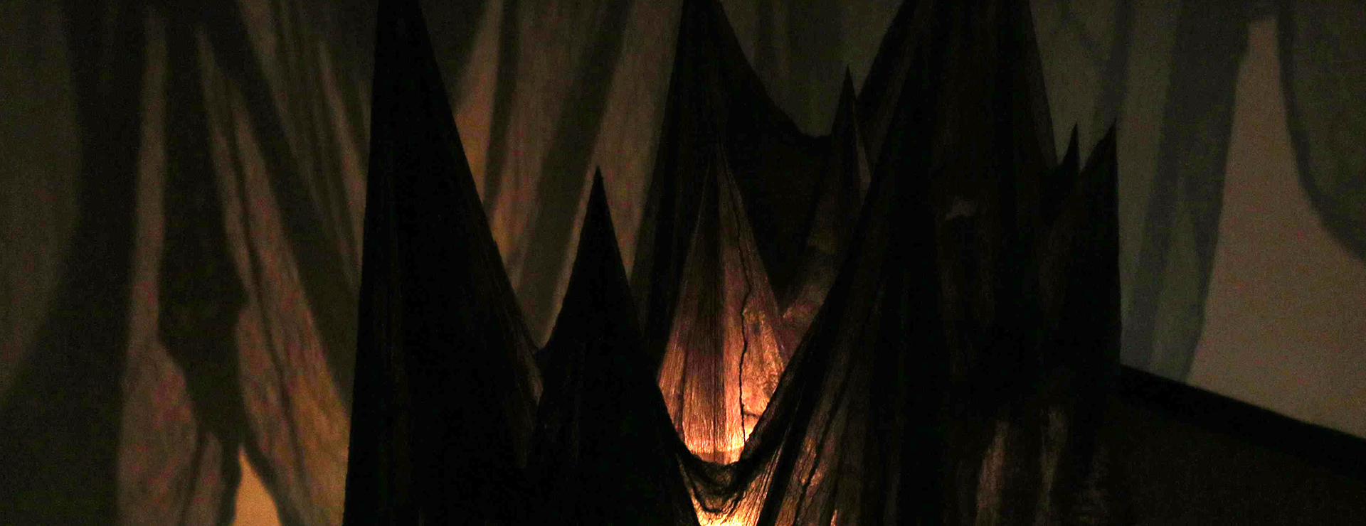 The image shows a cloth hanging from the ceiling, gently pulled downward by small sand pockets that create uneven folds and tension. A single light source shines at the center, illuminating the fabric while casting soft, shifting shadows on the surrounding walls, adding depth and a sense of quiet intensity.