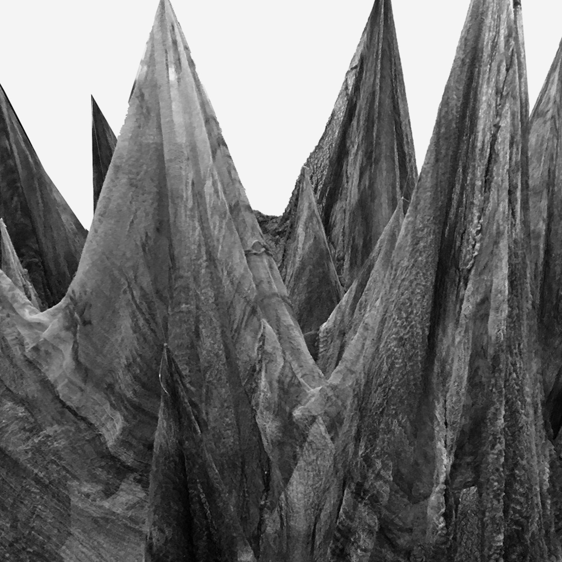 The black-and-white image shows a thin cloth suspended from the ceiling, its surface draping and folding into soft, uneven peaks and valleys. Without visible sand, the fabric’s contours resemble an abstract mountain range—light and shadow creating depth, texture, and a quiet, atmospheric sense of weight.