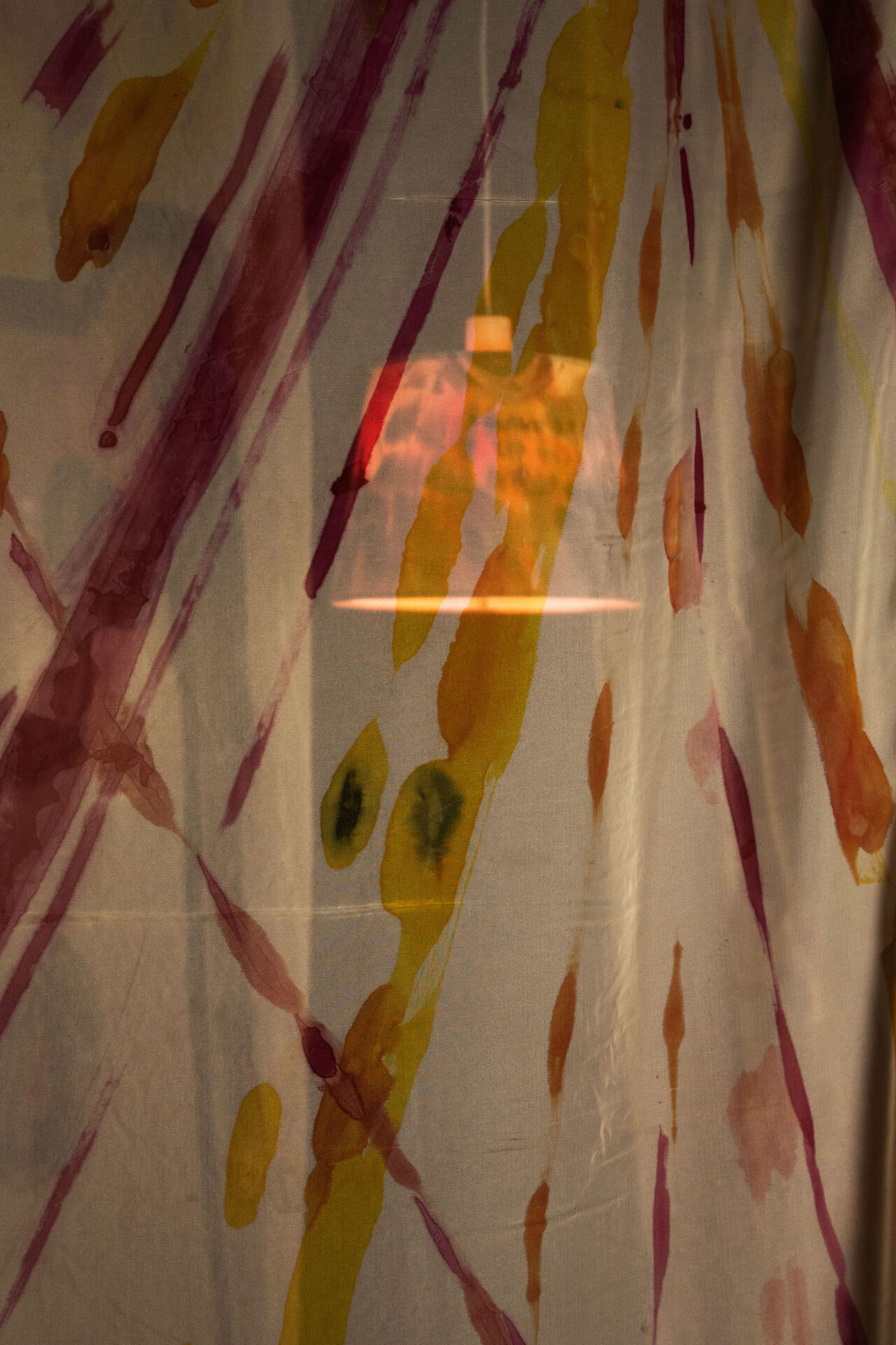 A black, yellow, and pink lamp shines behind a sheer curtain with streaks of colour painted on it