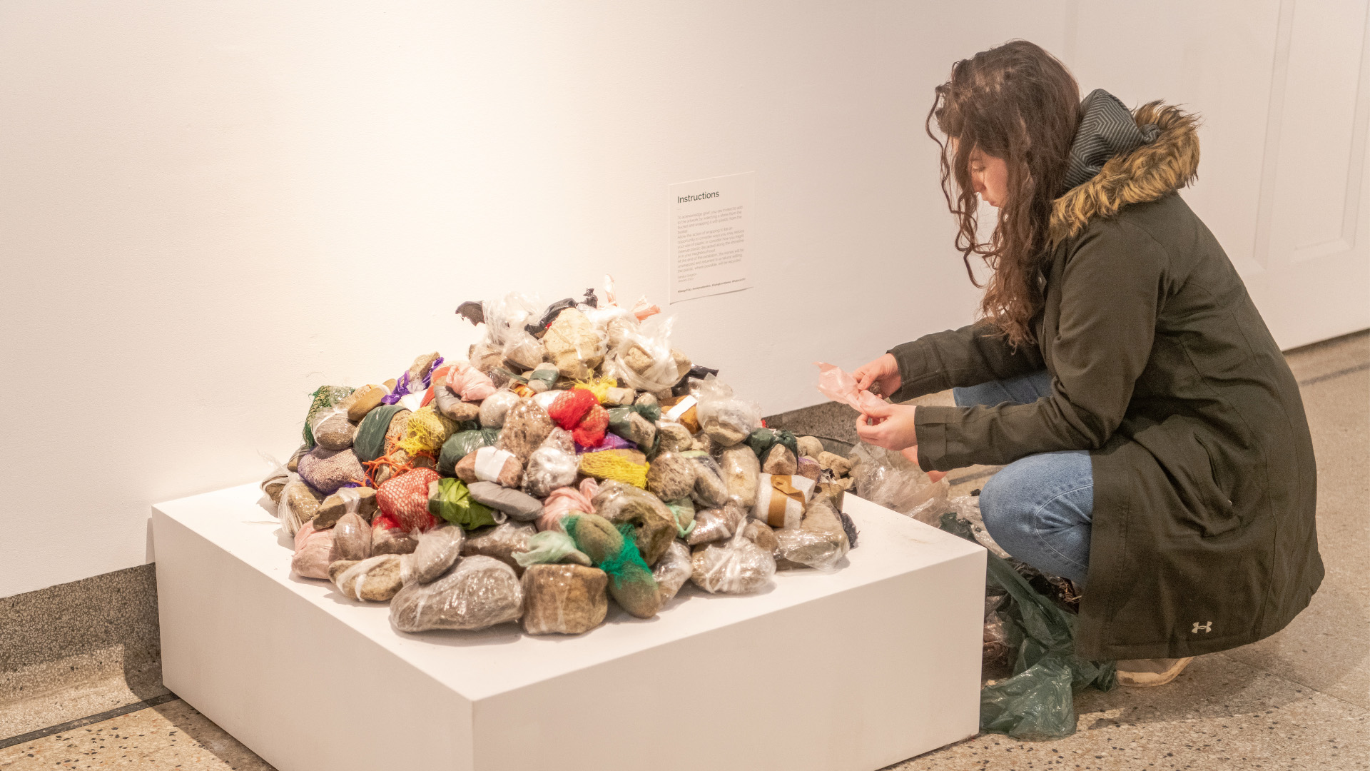 An attendee crouches down, carefully picking up a small, plastic-wrapped stone from a large pile of stones on a white pedestal. A sign with instructions is visible behind the pile.