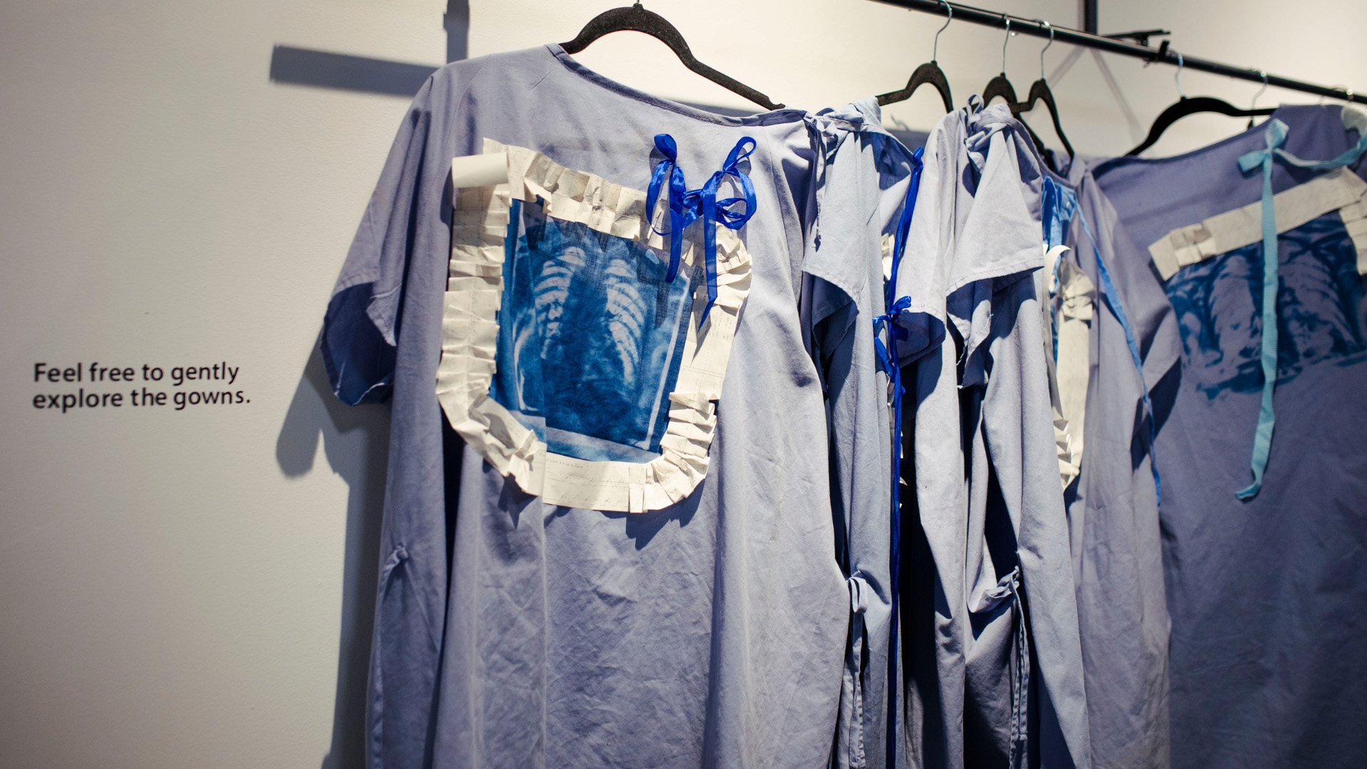 An installation features several light blue hospital gowns hanging on a rack. Each gown has a distinct rectangular patch with a blue and white cyanotype print of a human ribcage. A wall to the left has text that reads, "Feel free to gently explore the gowns."