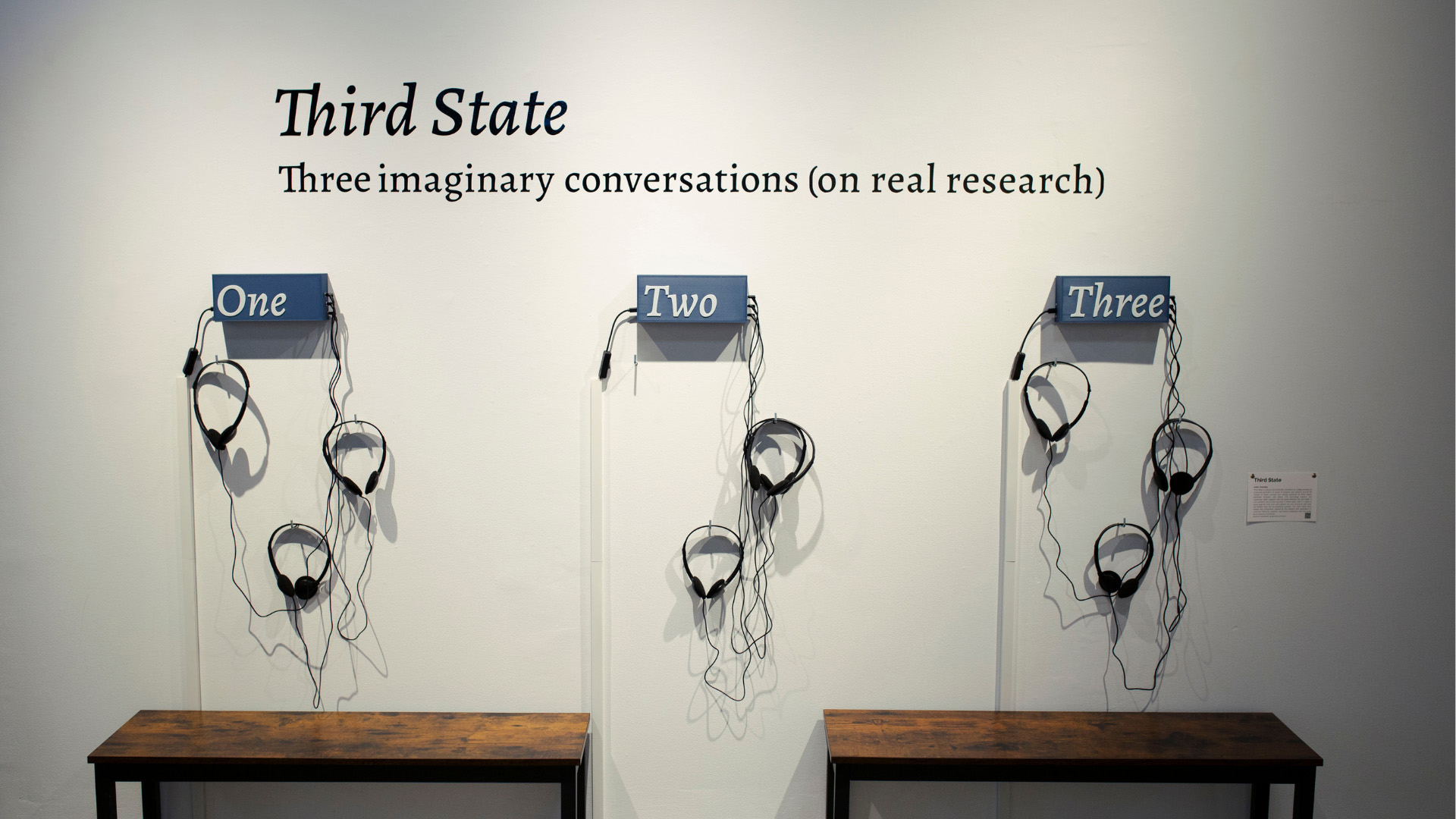 A gallery wall features three identical stations, each with a wooden bench, three sets of black headphones hanging from the wall, and a sign above. The signs read "One," "Two," and "Three," and the headphones are arranged as if for listeners.