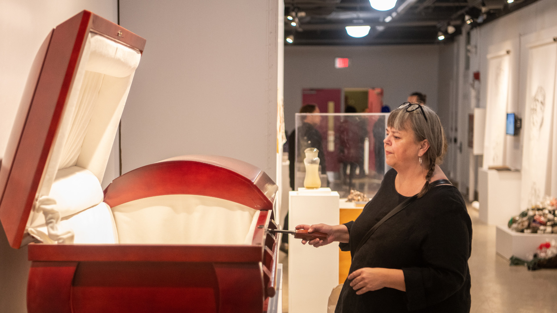 An attendee with her hair in braids and wearing a black shirt stands in a gallery, turning a small crank on an open, red coffin-shaped art piece. The crank is connected to an interior music box.