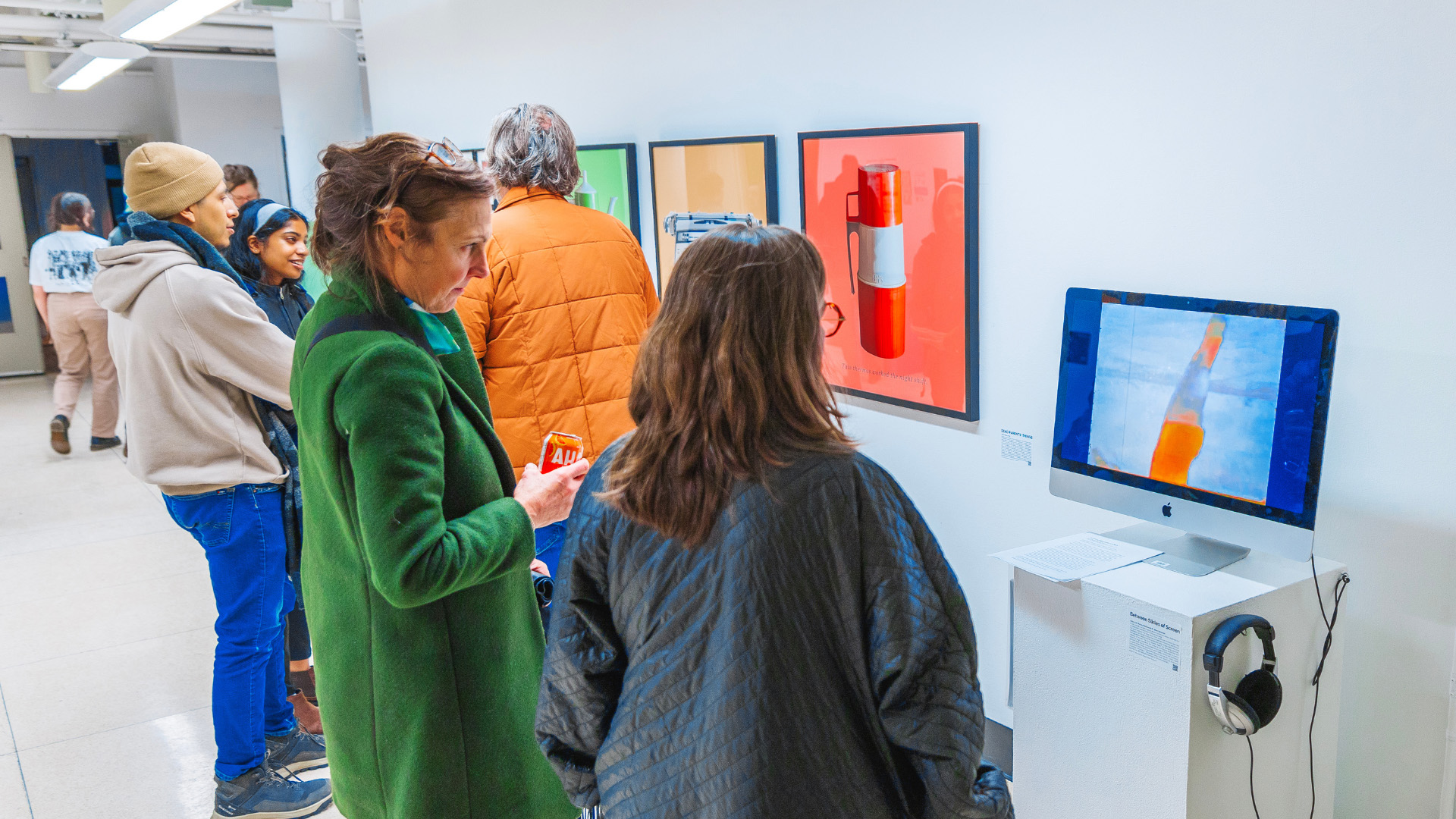 A group of people are gathered in a brightly lit gallery space, looking at various art pieces. One person in the foreground looks at a computer monitor displaying a bright orange and blue image. In the background, framed artworks are visible on the wall.
