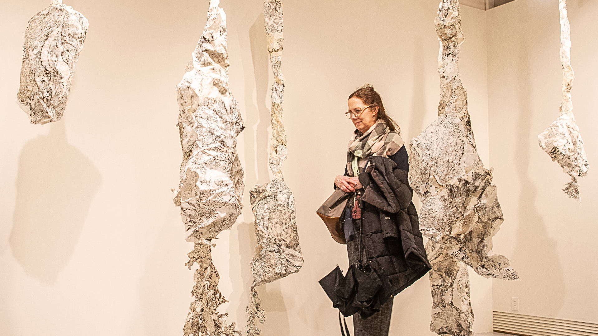 An attendee stands in a gallery, looking at several hanging art pieces. The pieces are collagraph prints of foraged lichen on rice paper, displayed as irregular, textured, white forms suspended from the ceiling.