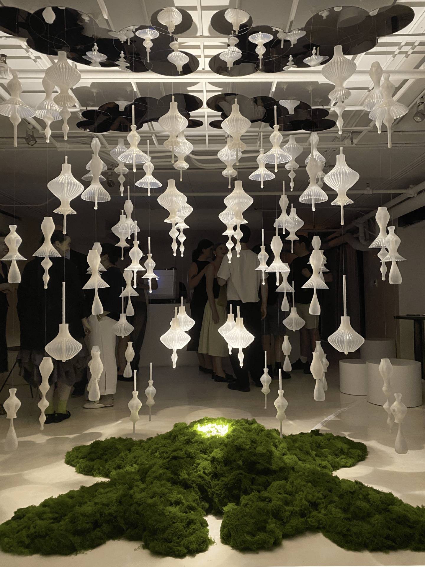 The same installation is shown from another angle, now with people standing and observing. The hanging ornaments shimmer under the light, while the green moss mound below anchors the piece like a living landscape. The visitors’ presence emphasizes the scale and immersive quality of the artwork—inviting reflection on how nature, architecture, and human connection intertwine within the space.
