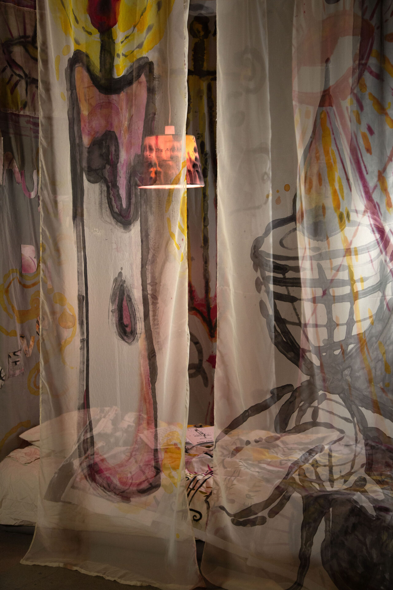 A black, yellow, and pink lamp shines over a bed behind two sheer curtains. One has a chalice painted on it, and the other has a candle painted on it