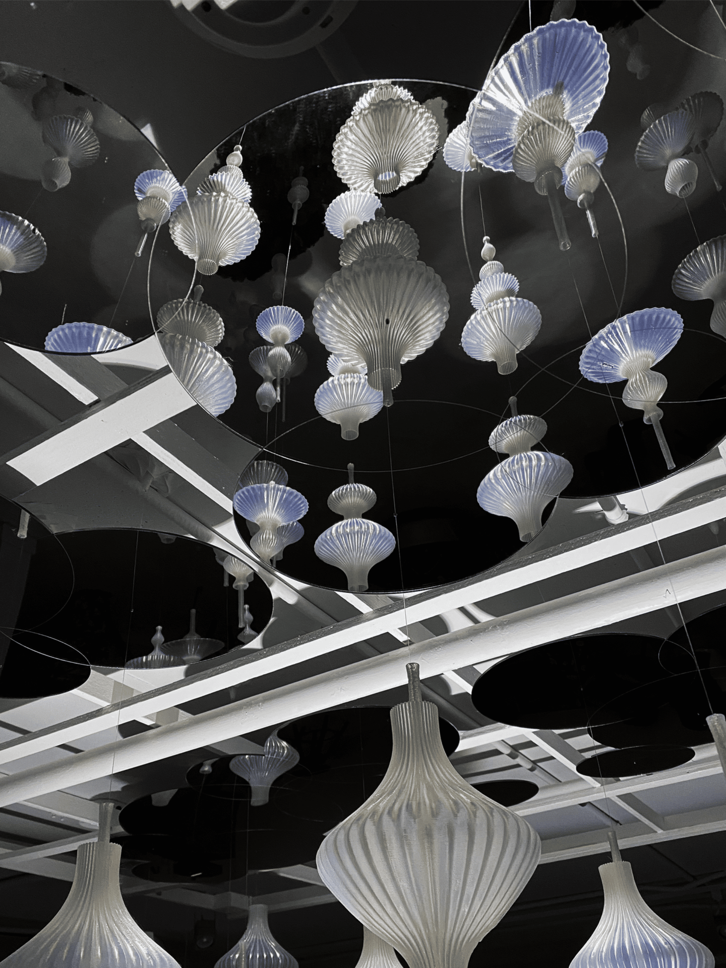 The image shows a ceiling installation composed of multiple hanging translucent ornaments shaped like delicate shells or fluted lanterns. The translucent forms glow softly in shades of white and pale blue. Above them, circular mirrors reflect the hanging pieces, creating an illusion of infinite depth and floating repetition. The structure holding them is made of white intersecting beams, giving the scene an airy and architectural feel.