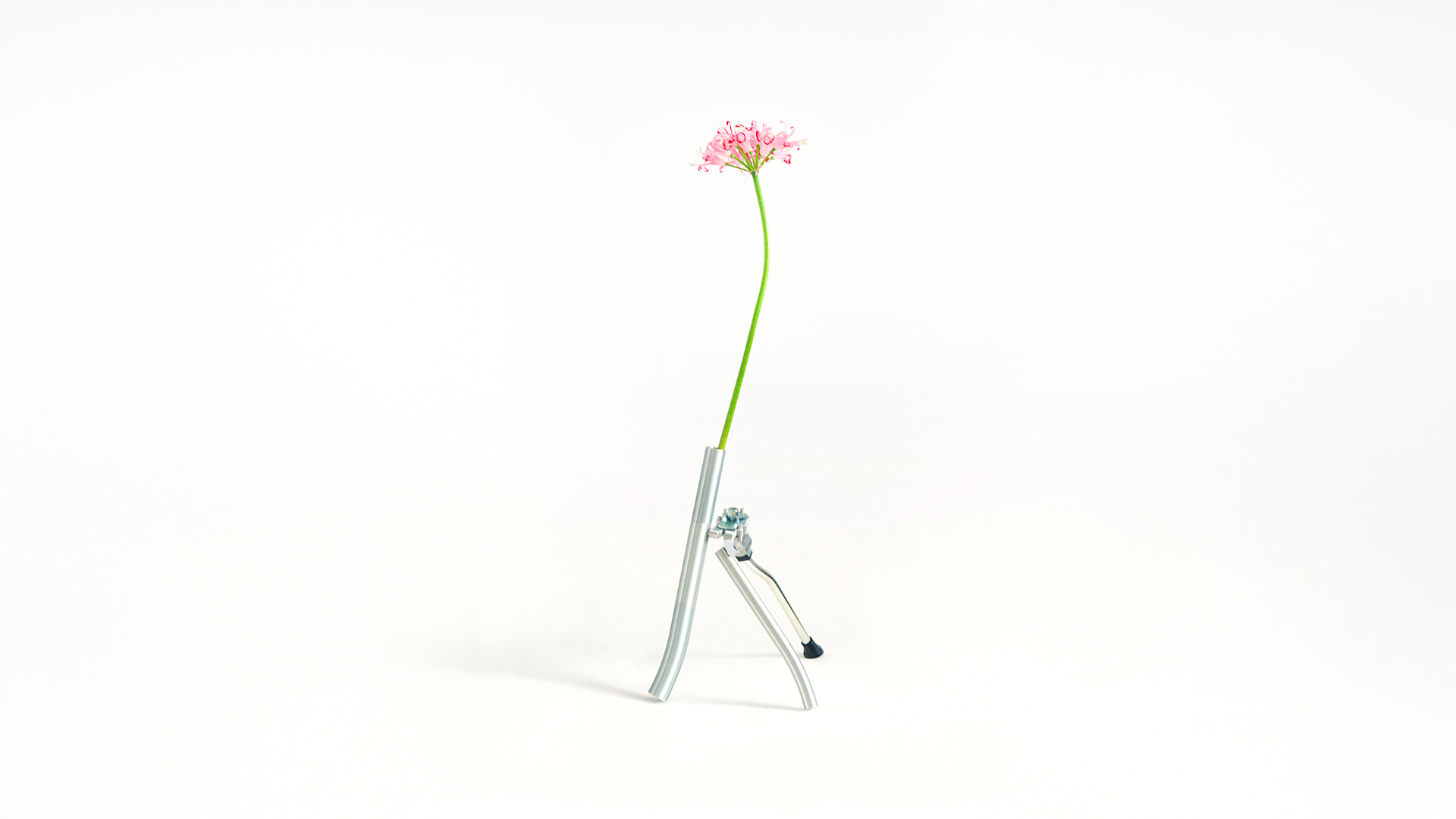 A minimalist studio photograph of a sculptural vase made from silver 3d printed tubes and a bicycle kickstand component, holding a single pink flower with a long green stem.