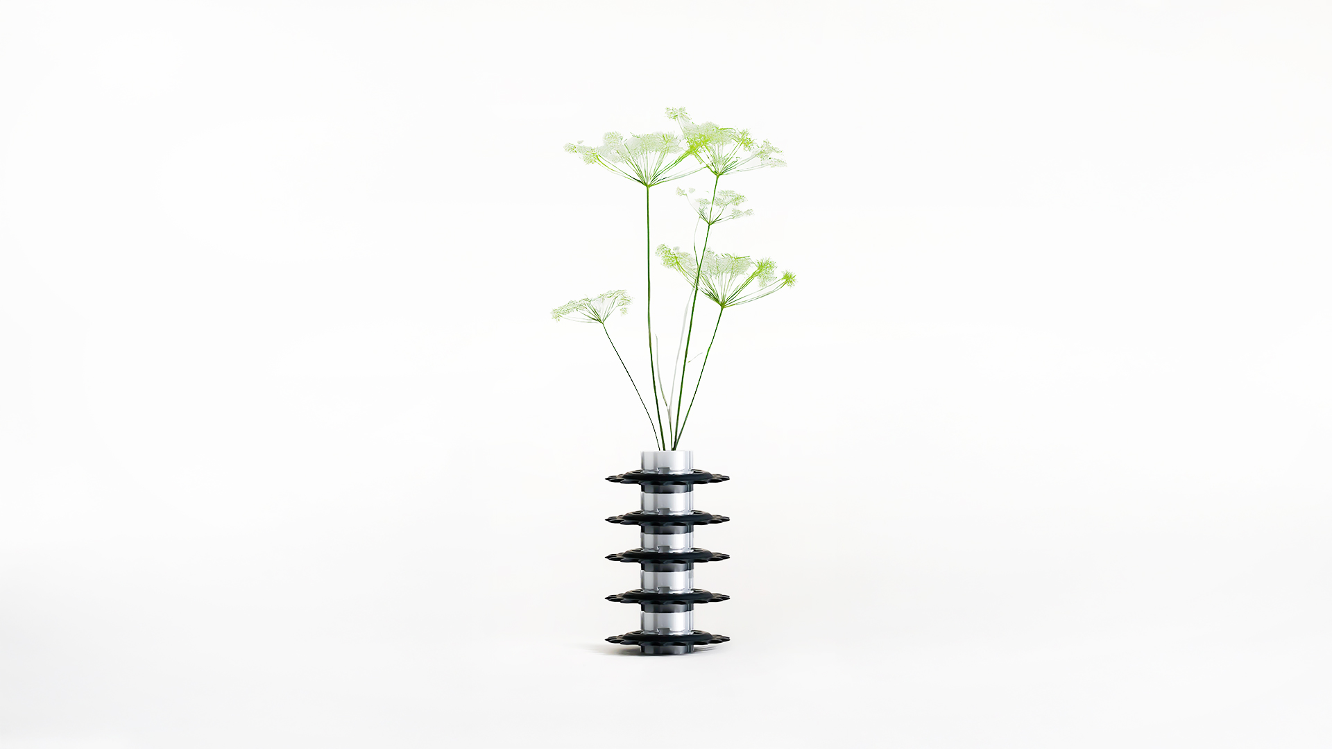 A minimalist studio photograph of a sculptural vase composed of stacked black bicycle sprockets and silver 3d printed cylinders, holding several delicate green stems.