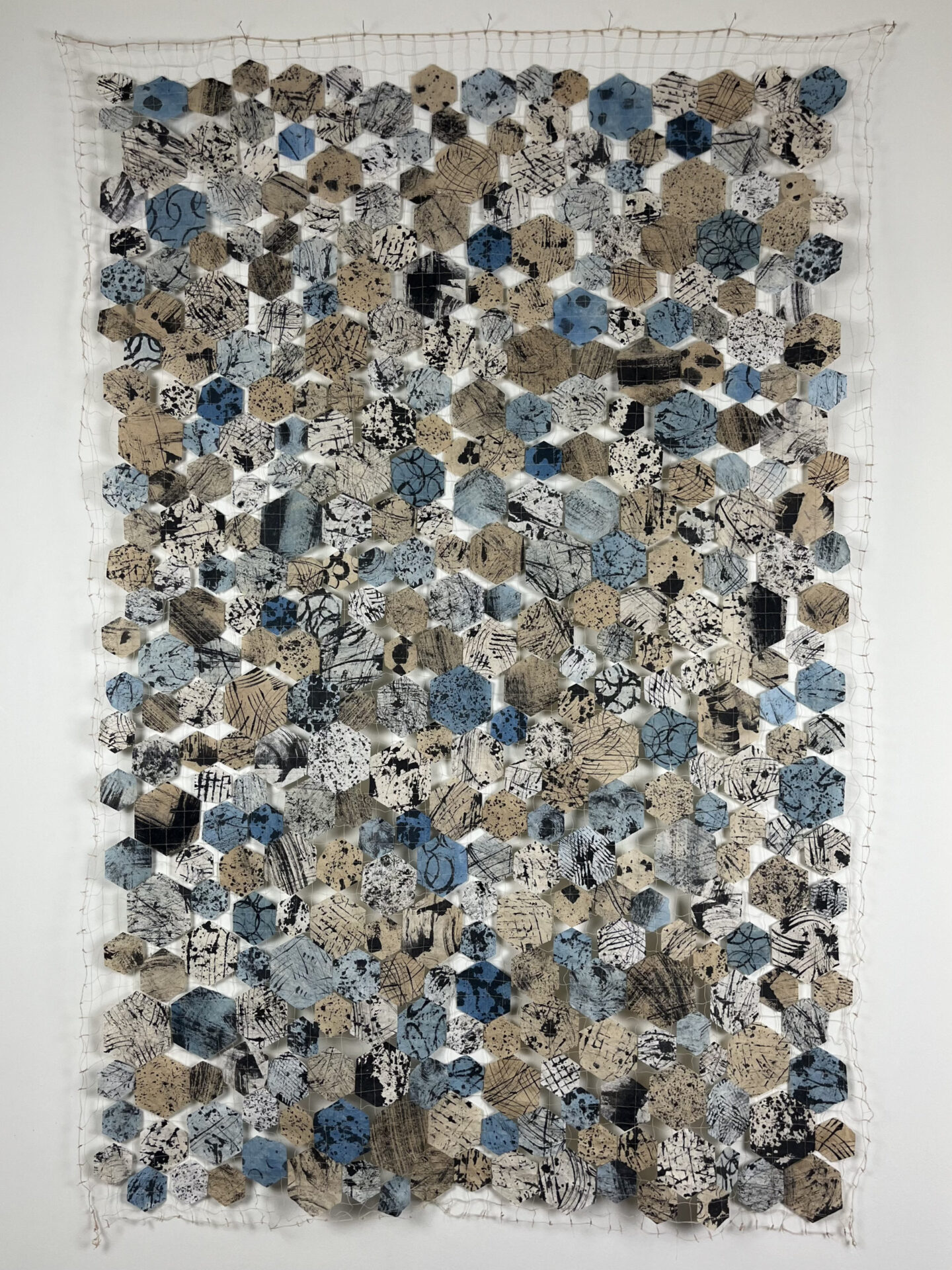 "Flow", hexagonal fragments of used coffee filters, patterned with ink and dyed with indigo or tea, and sewn into a random pattern on an open thread grid, forming a quilt-like panel.
