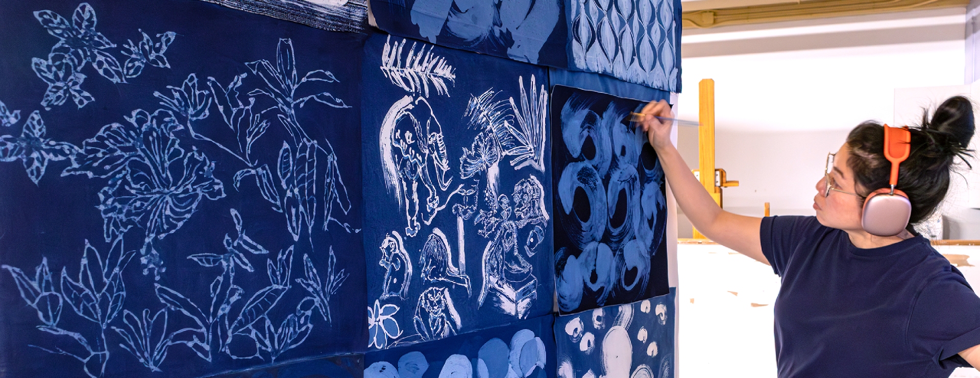 The artist, wearing earphones, stands next to indigo batik samples with a brush resting on the fabric.