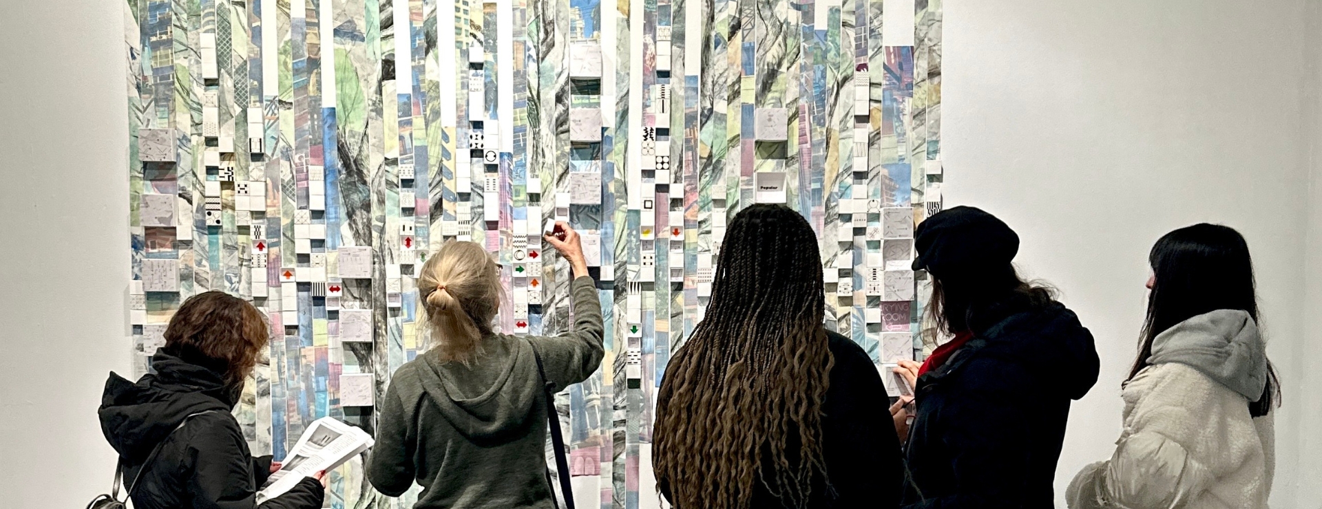 An installation photo showing a group of five people interacting with a large-scale art installation made up of small paper boxes that can be re-arranged. Each box shows a different symbol, arrow, or pattern  according to different elements found in cities. 