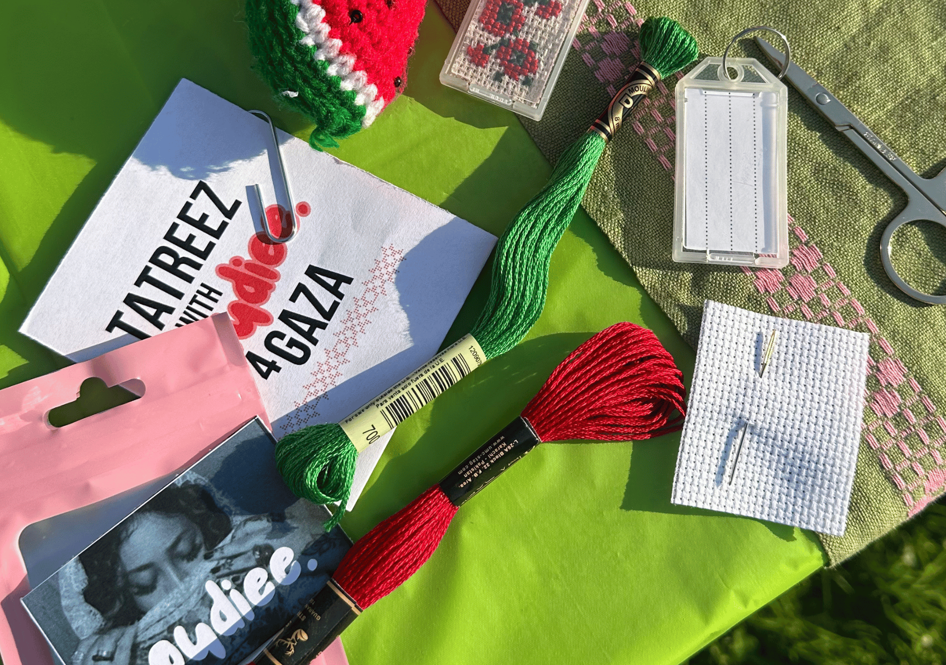 A bright, close-up photograph captures materials for a Palestinian embroidery (Tatreez) activity laid out on a lime green surface in sunlight. The image includes red and green embroidery threads, a small piece of white fabric with two needles, silver embroidery scissors, and a woven green cloth accented with pink stitching.
A card in the center reads “TATREEZ with noudiee. 4 GAZA”, connecting the scene to a community art initiative.