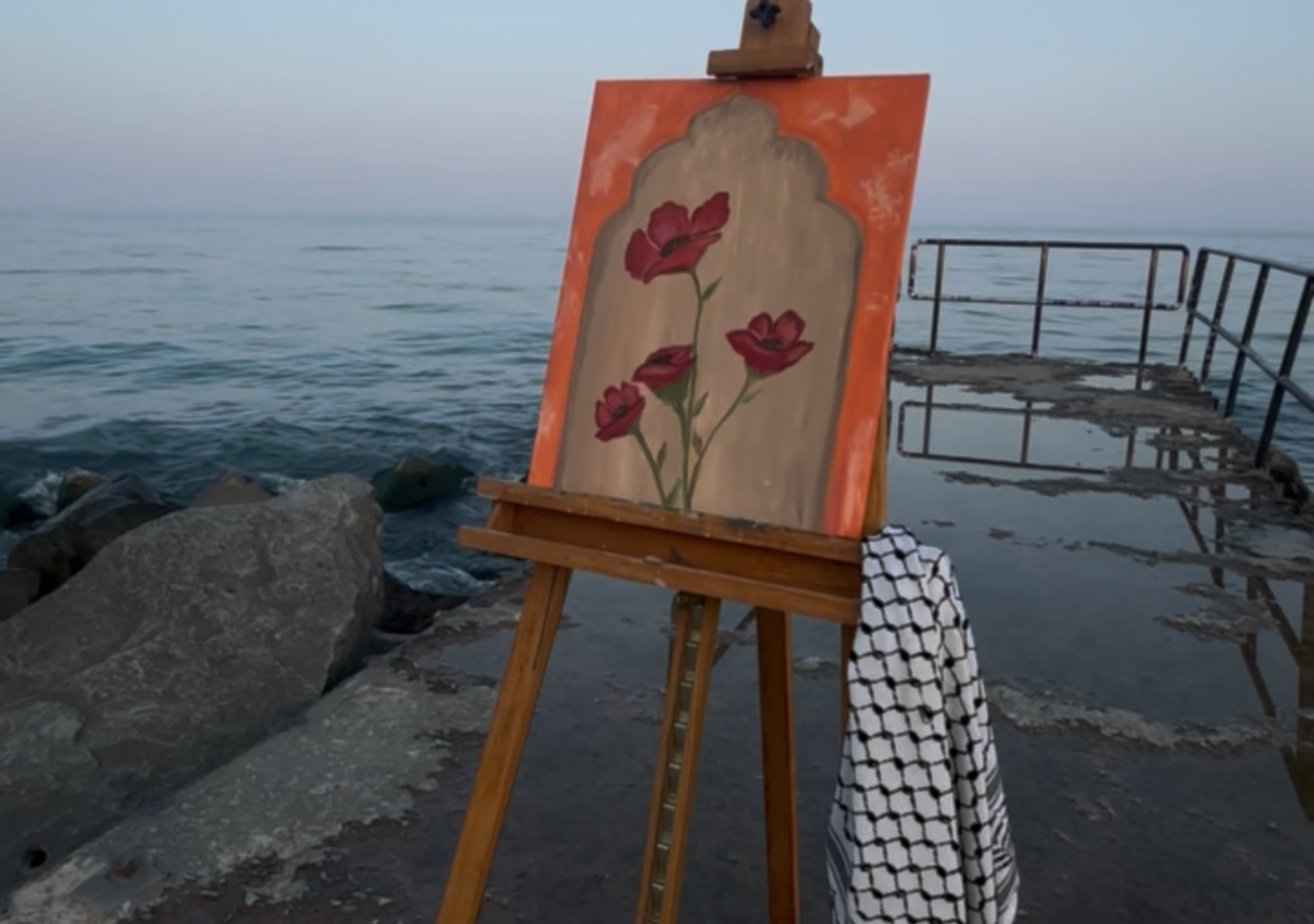 A wooden easel stands at the edge of a pier, facing the calm sea beneath a soft blue sky. Resting on it is a painting framed by an arched doorway — a motif drawn from traditional Palestinian architecture, serving as a window to home and heritage. Within the arch, red poppies bloom, symbols of memory, resilience, and belonging. Draped over the easel’s leg, a black-and-white keffiyeh anchors the scene in identity and resistance.