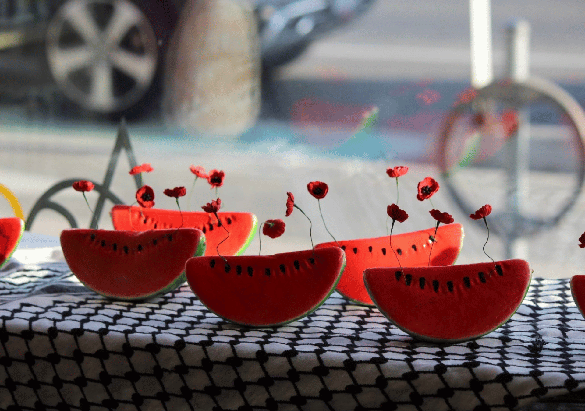 Watermelon slices arranged on a keffiyeh-patterned cloth, each with delicate red poppies sprouting from the top.