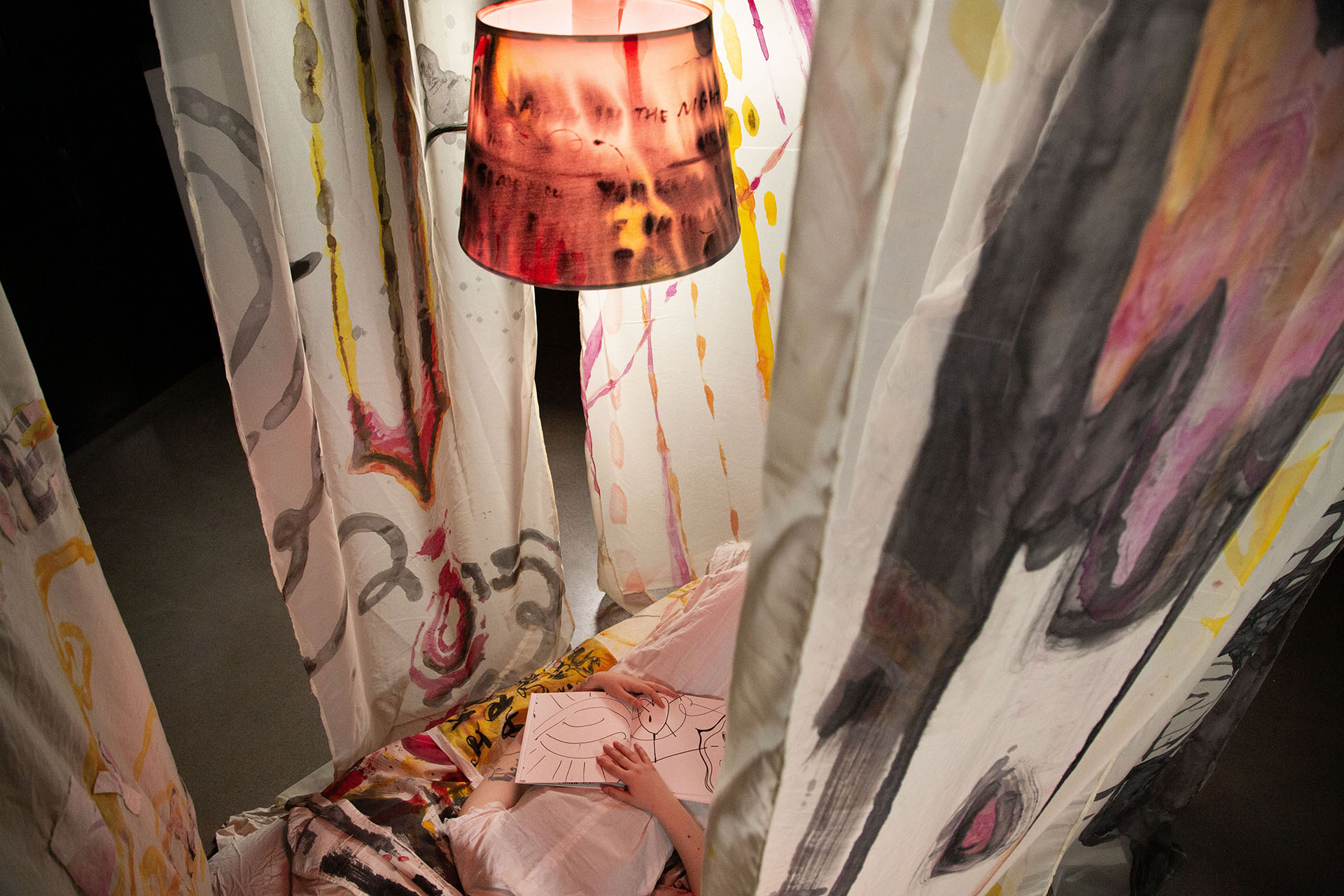 The torso of a person in a white dress lies on a bed with a printed quilt underneath is shown from above, next to their hand is an open book with drawings in it, in the frame is a black, yellow, and pink lamp 