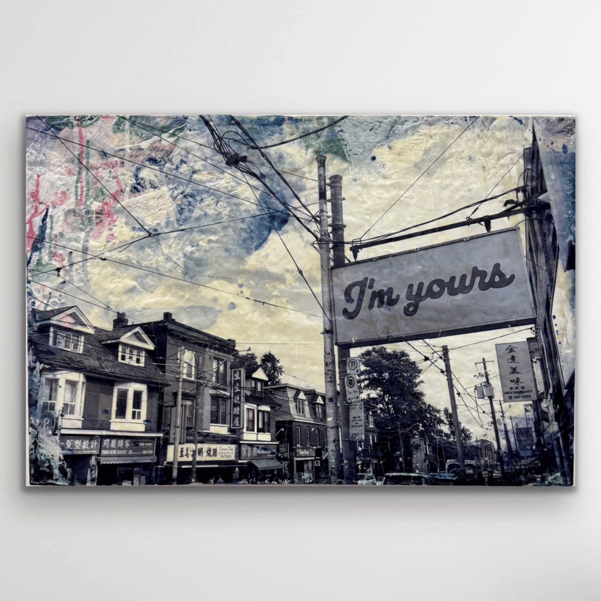 Black and white photograph of the text 
"I'm Yours" hanging from a sign with a row of urban Toronto buildings in the background. Spatters of paint and encaustic wax surround the image. 
