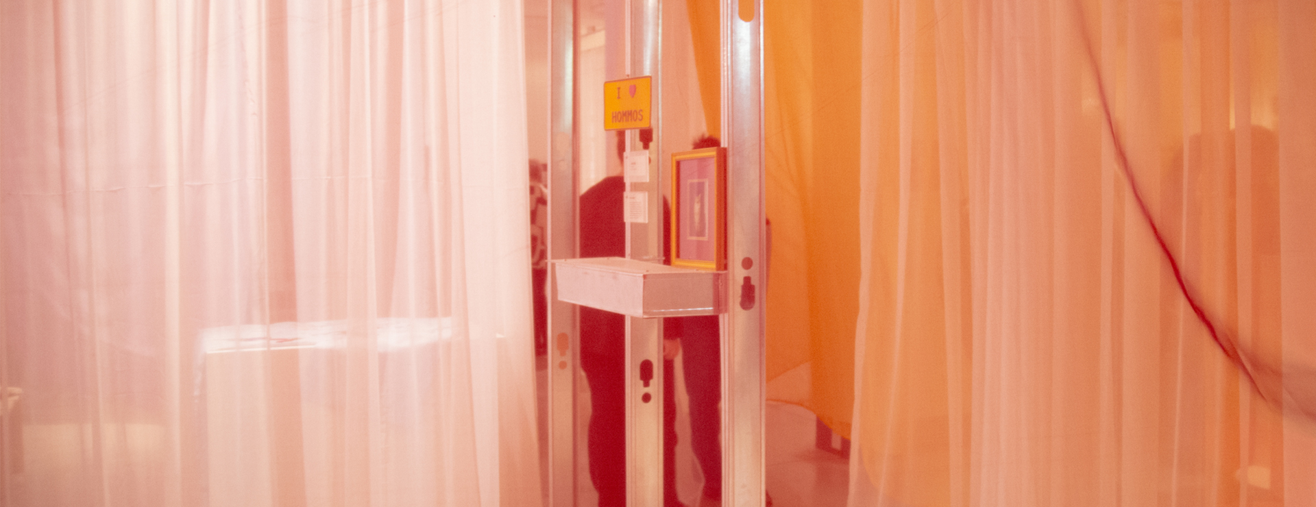 A soft, ambient image of the TO ·BE·LONGING exhibition space. Sheer, colorful rippled curtains create curved, translucent walls in shades of pink, yellow, and red. The lighting casts vivid magenta and blue shadows on the floor. The overall scene suggests a private, colorful, and intimate environment.