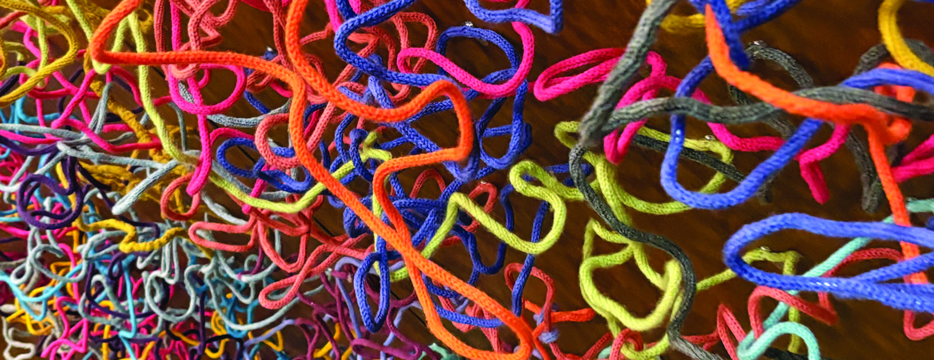 Sprawling soft sculpture made of knitted and textured yarn cords (orange, green, light blue, mustard yellow) on a dark background.