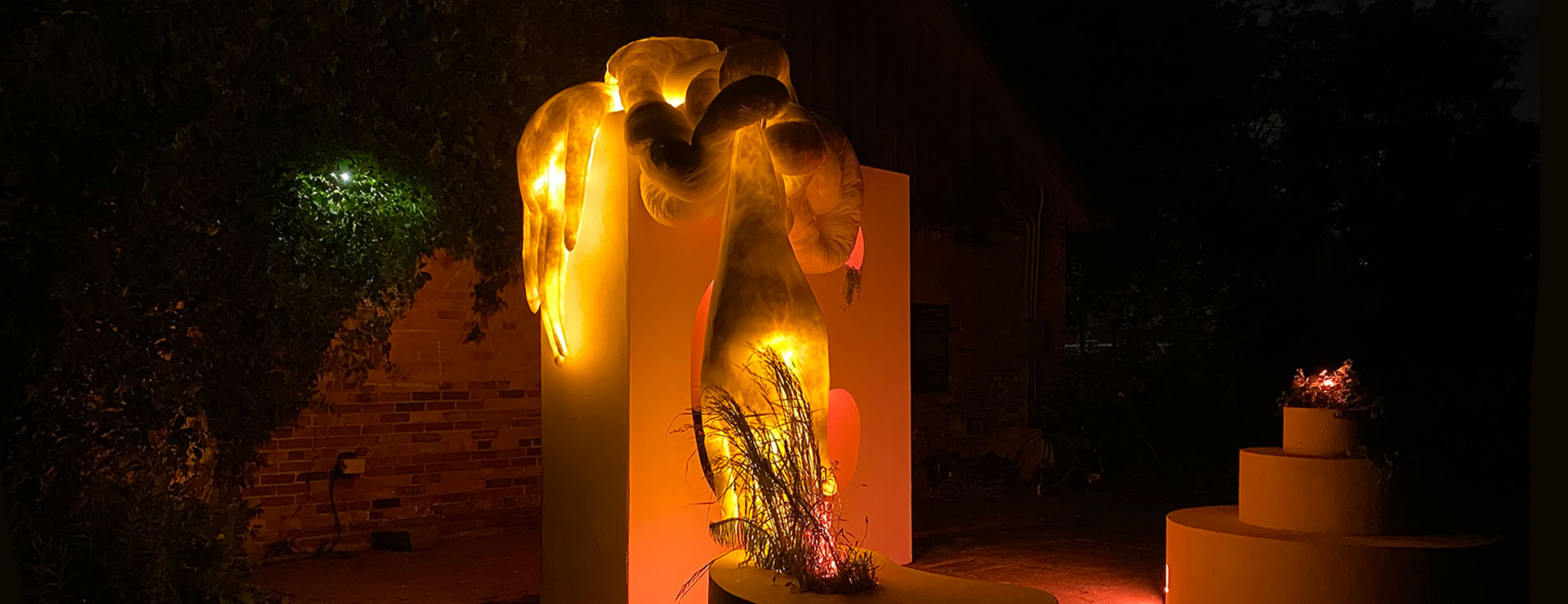 Intertwined soft sculpture in the form of long arms with plants glowing with orange light