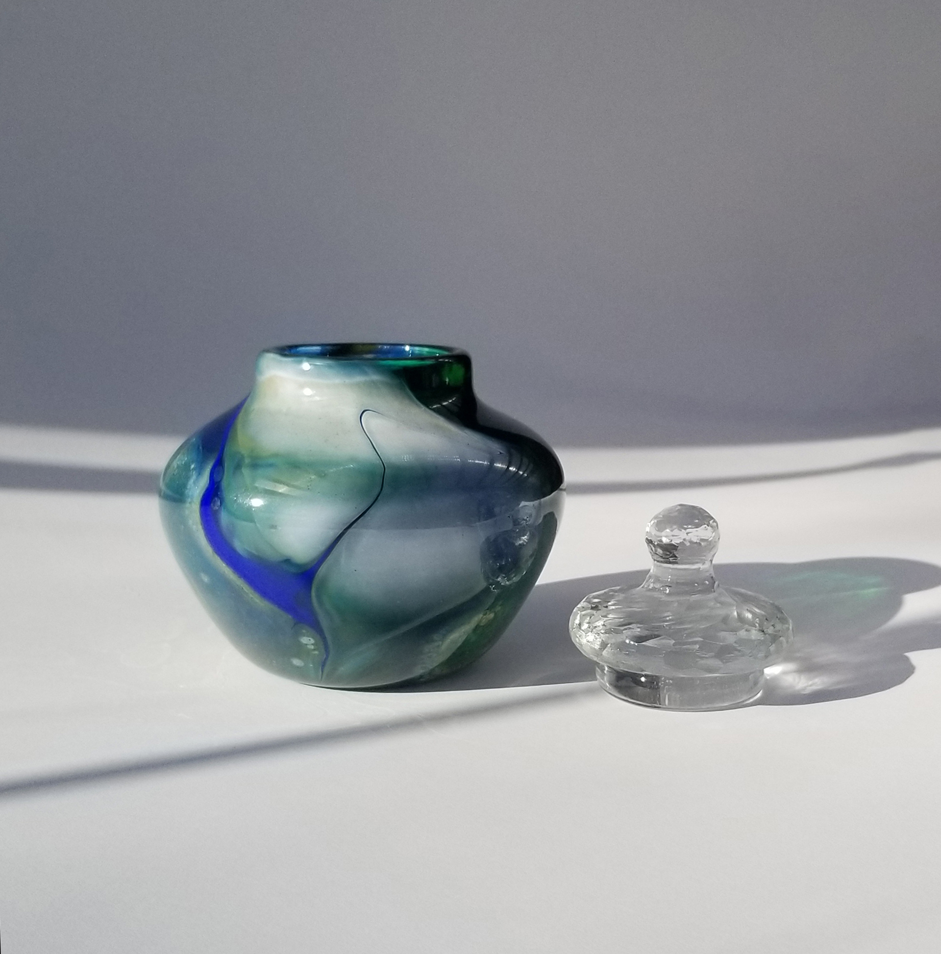 Image of a squat blown glass vessel in shades of blue, green and white against a light grey background. The vessel has a bold blue river running across the pale green surface. To the right sits a clear glass lid that has been carved like lead crystal.